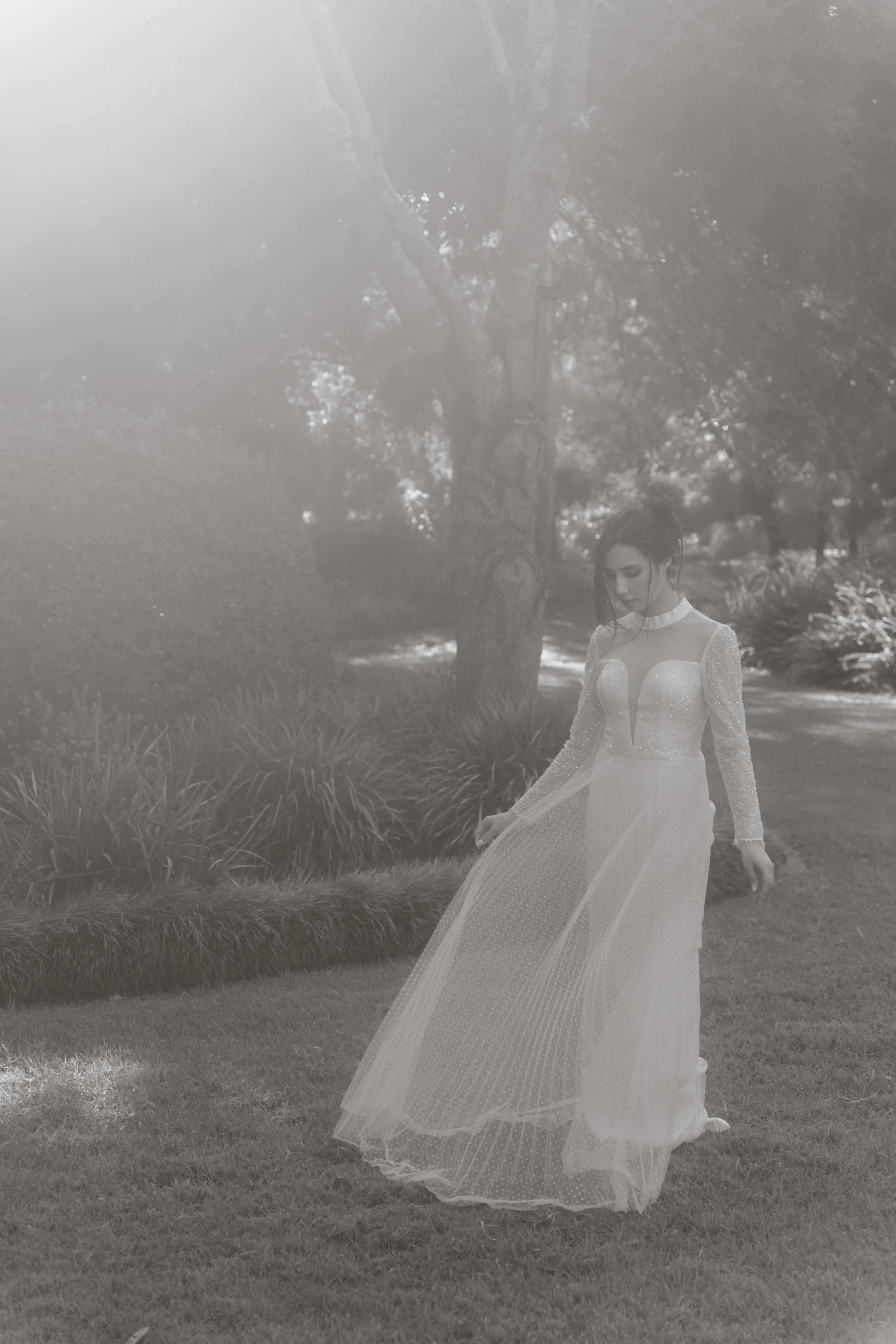A woman in a long, elegant dress standing on a grassy area in a park with trees in the background, the image has a soft, glowing light effect.