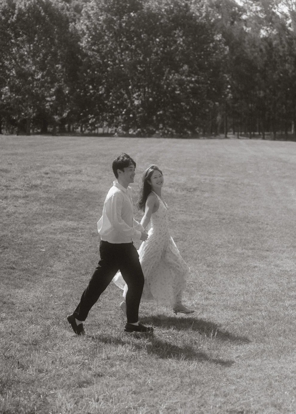 A black and white photo of a smiling man and woman holding hands and walking through a park on a sunny day, with large trees in the background.