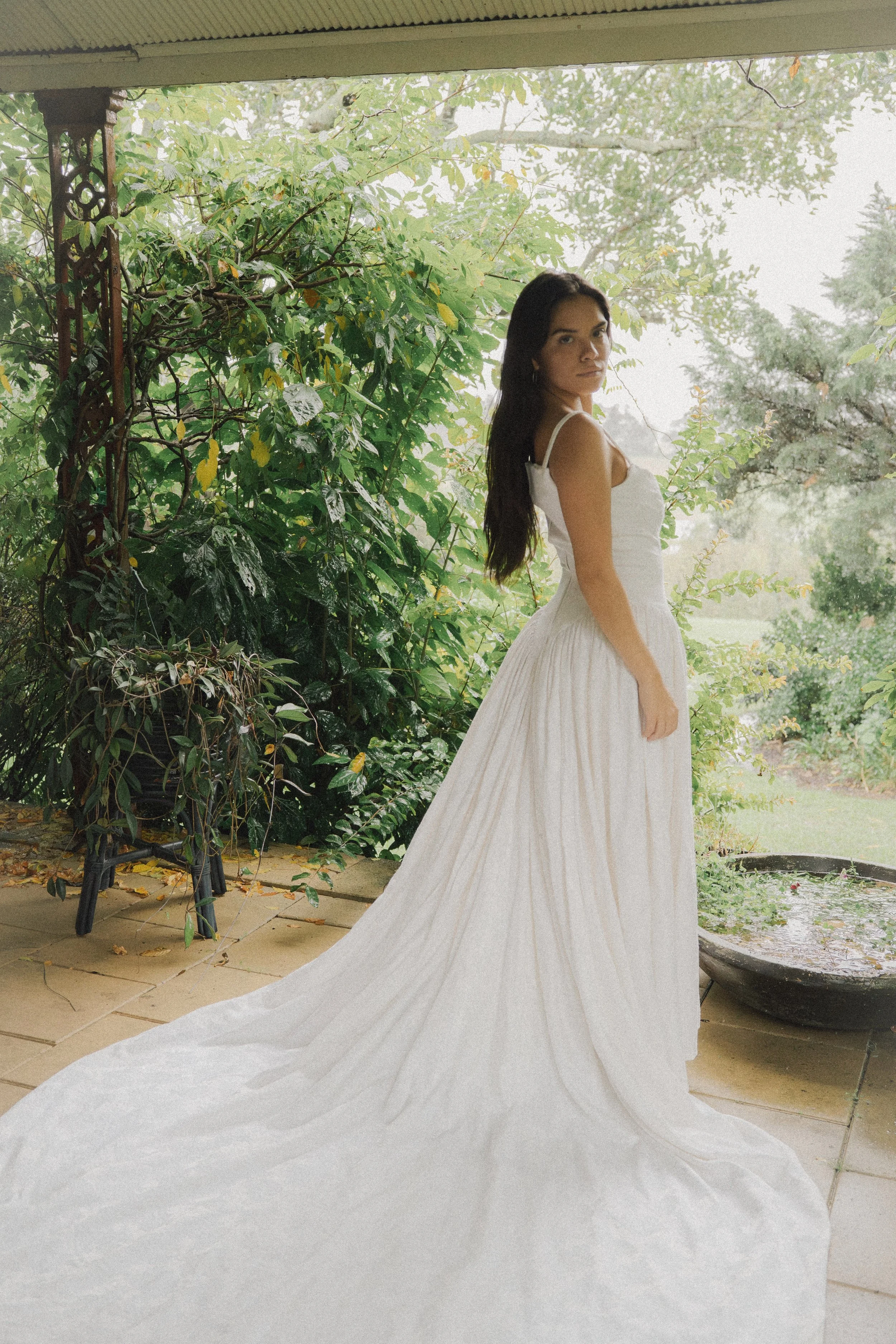 A woman in a white gown standing on a porch with lush green foliage and a garden in the background.