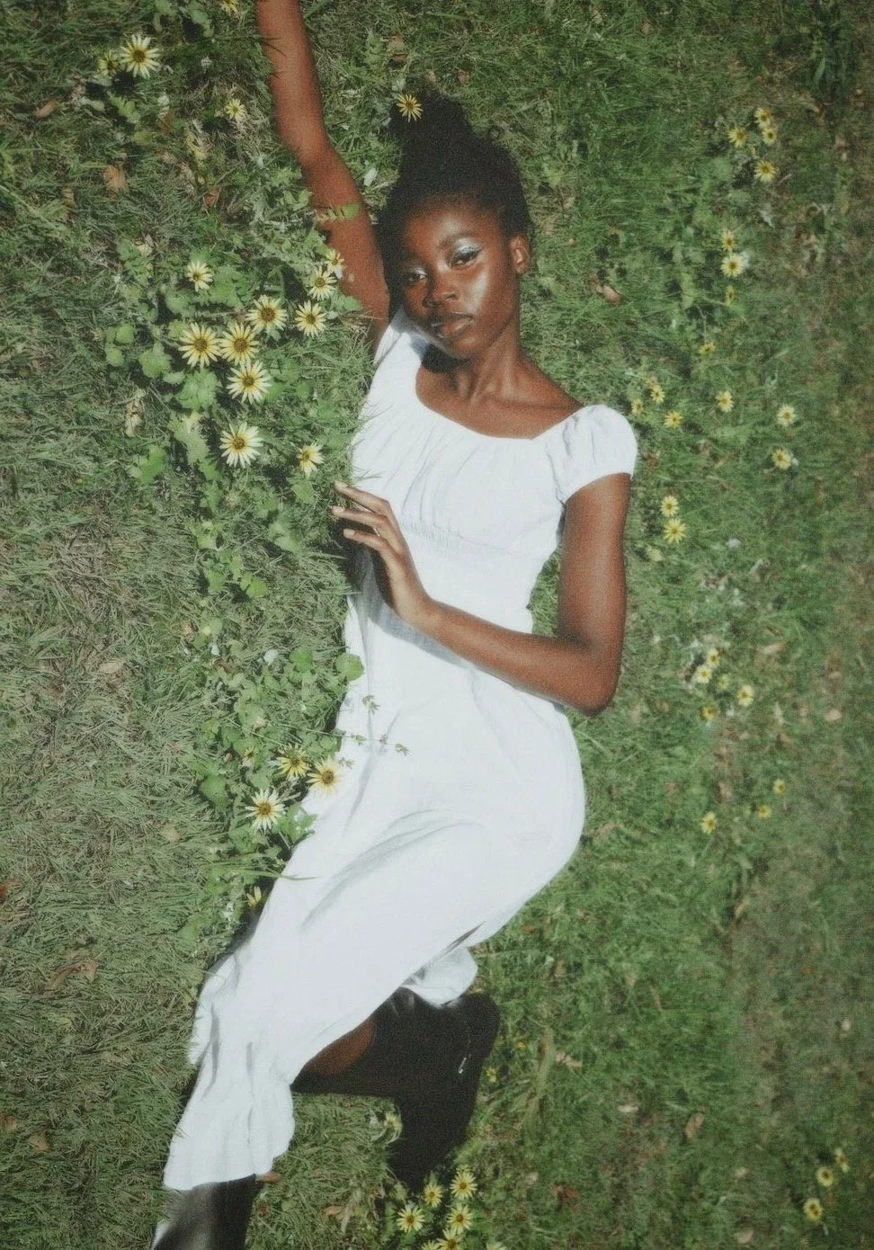 A woman lying on grass surrounded by yellow and white daisies, wearing a white dress and black boots.