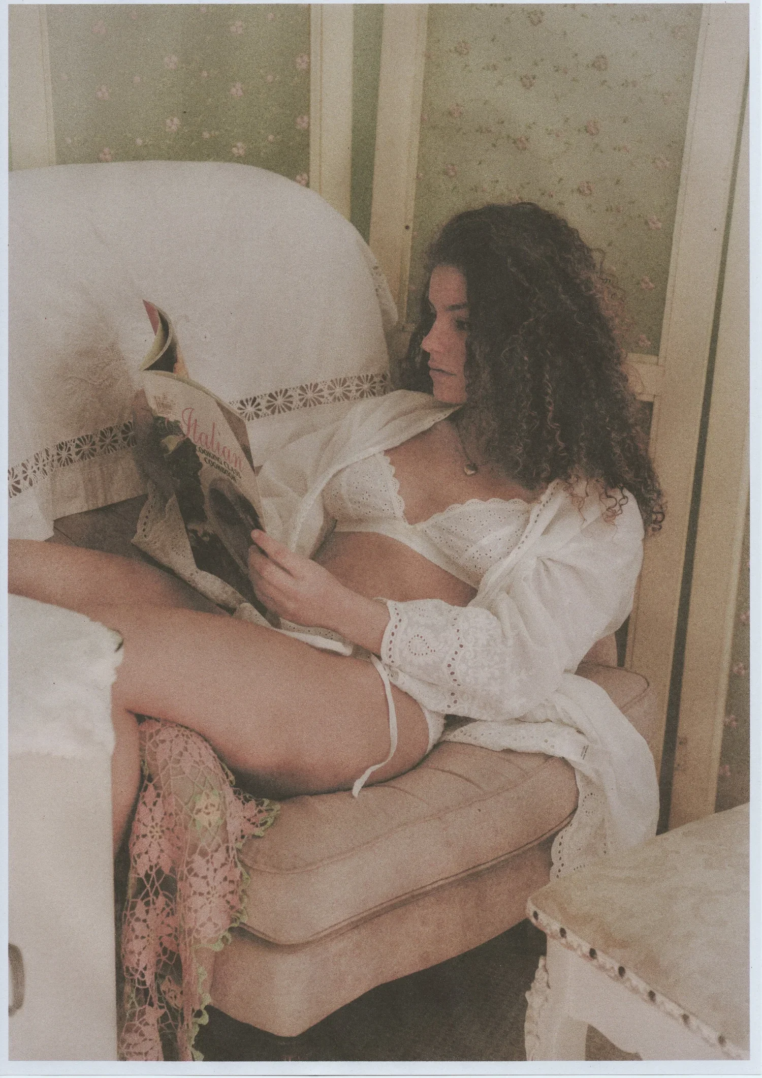 A woman with curly hair reading a magazine while sitting on a bed in a vintage-style room.