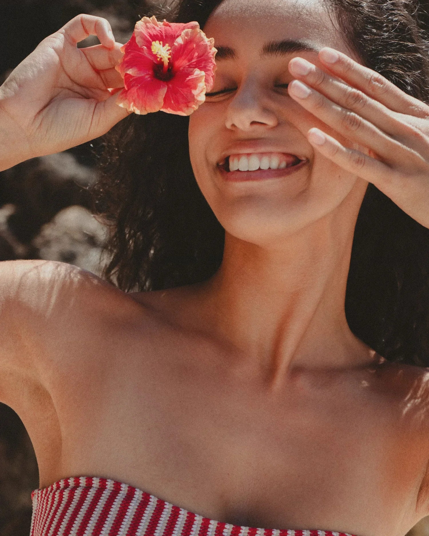 A woman smiling with her eyes closed, holding a pink flower to her face, wearing a strapless red and white striped top.