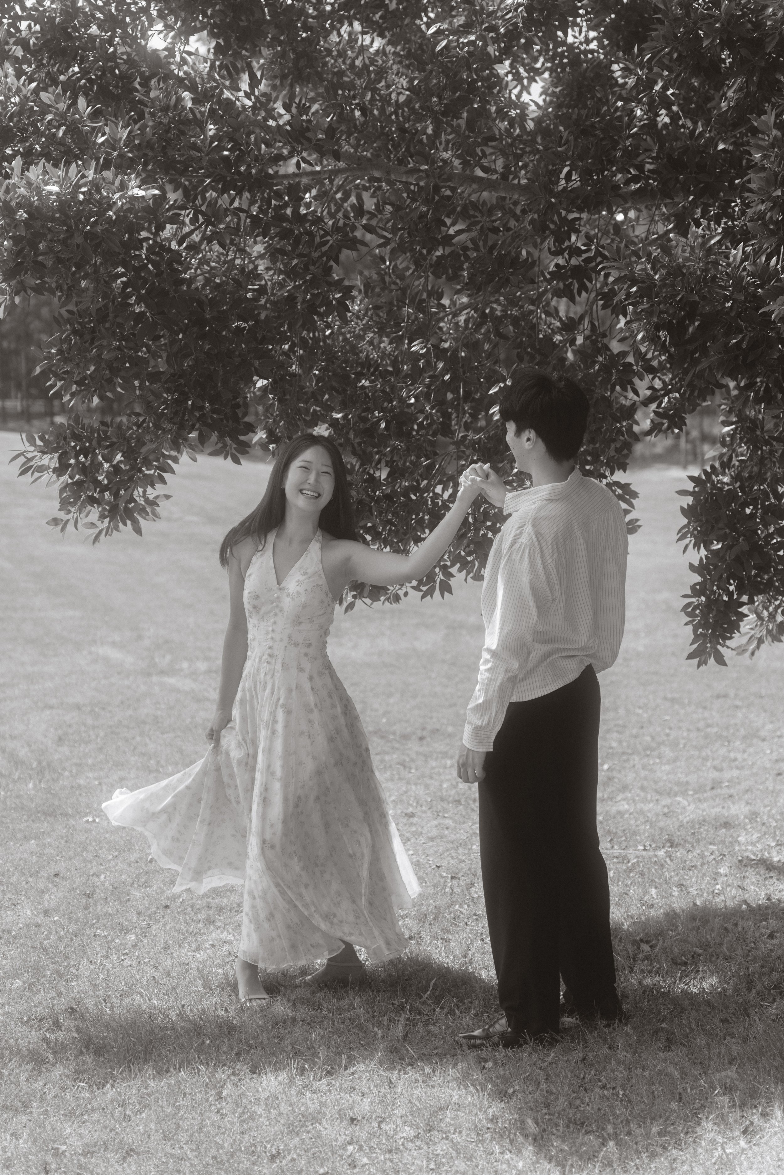 A woman and a man dancing outdoors under a tree, smiling and holding hands, in a black and white photo.