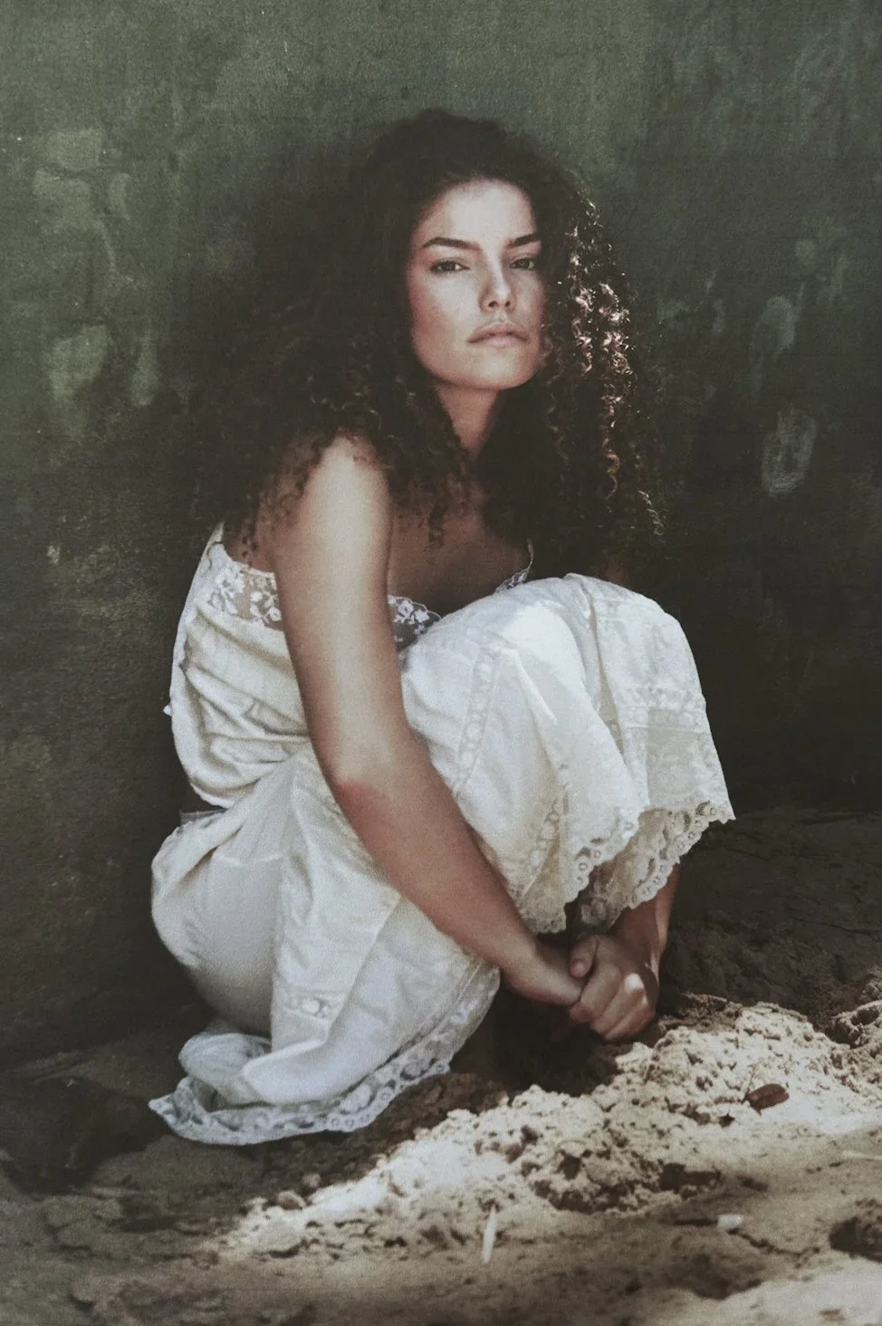 A woman with curly hair wearing a white dress, sitting on the ground with her knees up and hands clasped in front of her, against a dark, textured wall.