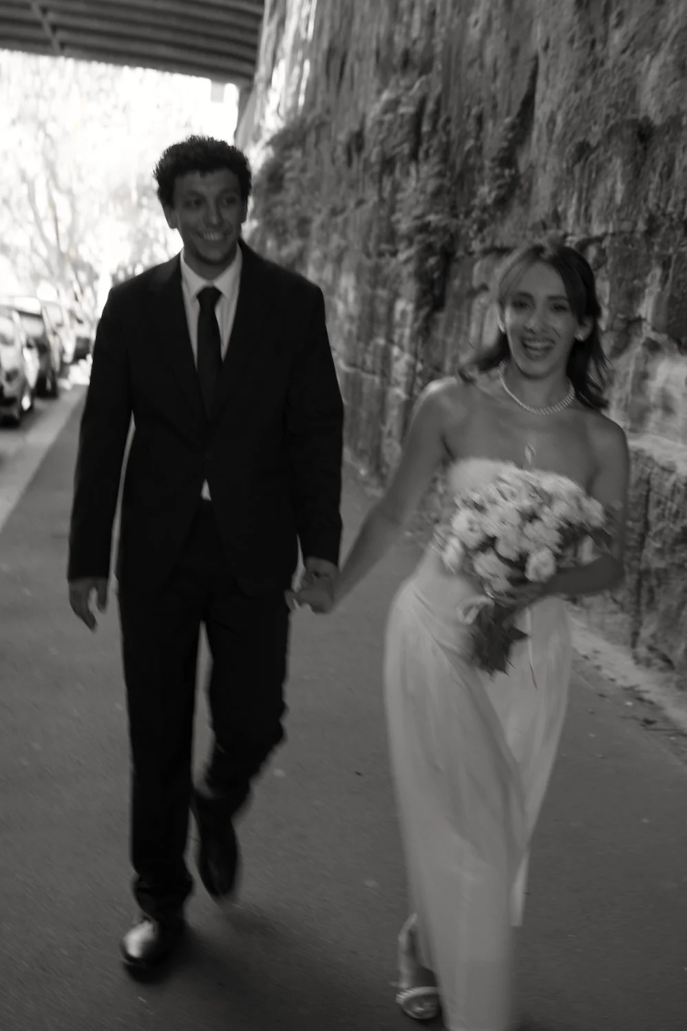A newlywed couple walking hand in hand along a street, smiling, with the groom in a suit and the bride in a wedding dress holding a bouquet of flowers.