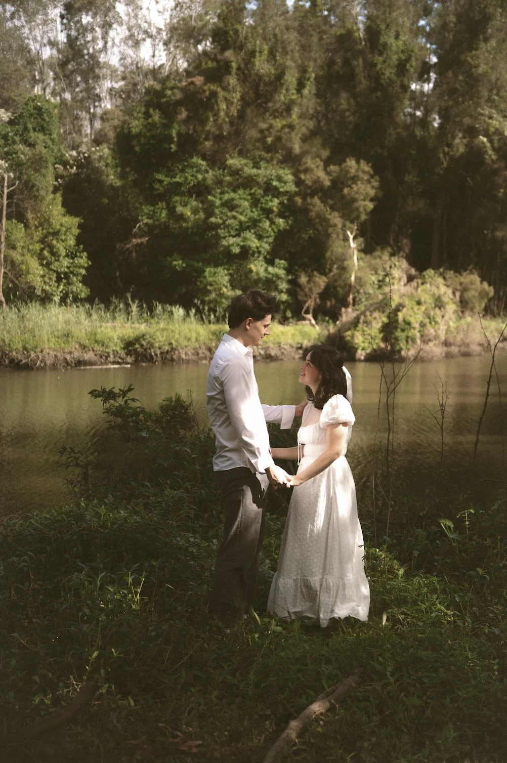 A couple in wedding attire standing by a lake, holding hands and looking at each other, with trees and greenery in the background.