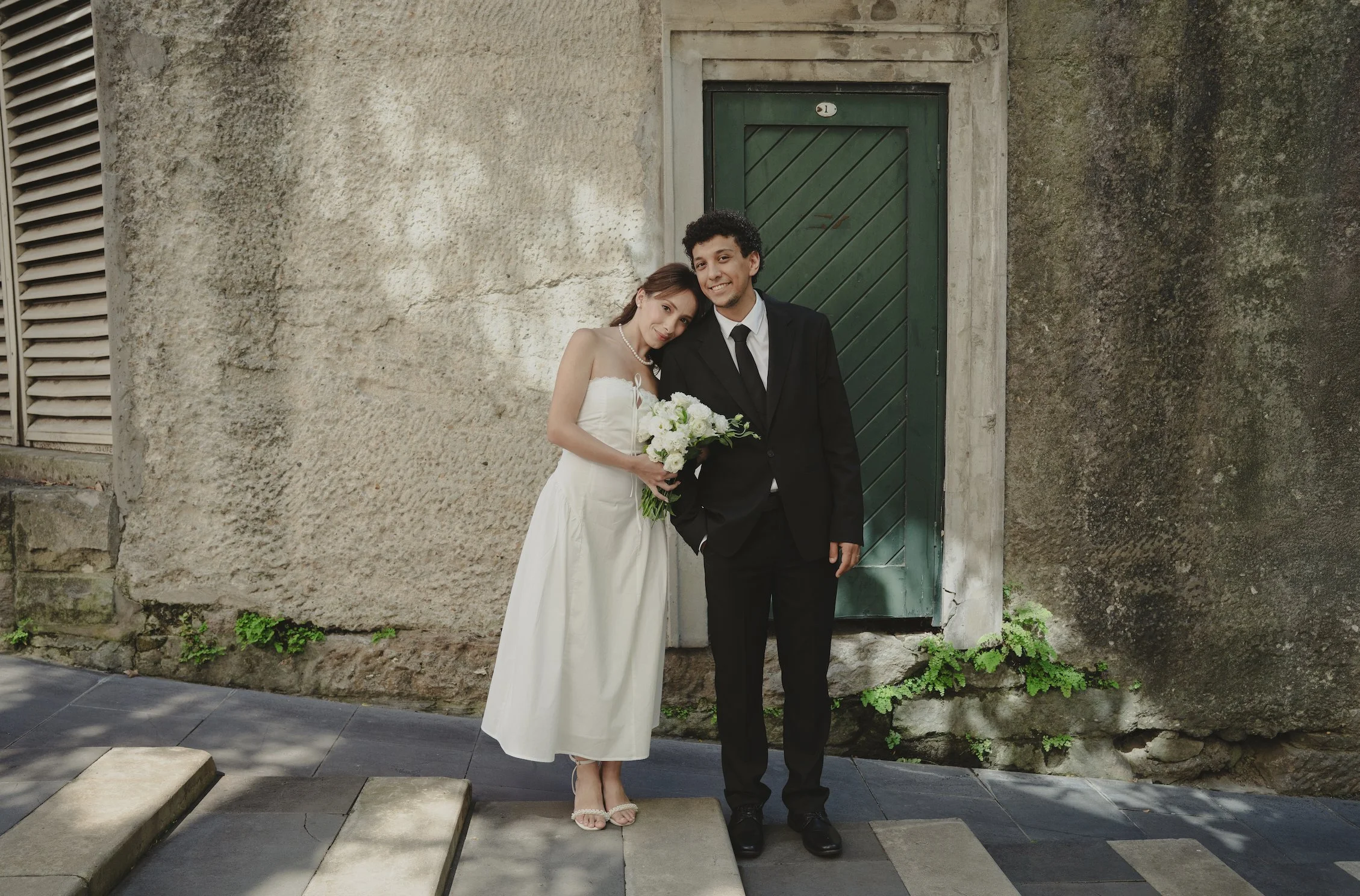 A bride and groom standing together on a sidewalk in front of a rustic wall with a green door. The bride is in a white wedding dress holding a bouquet, leaning on the groom's shoulder, and smiling. The groom is in a black suit with a white shirt and 