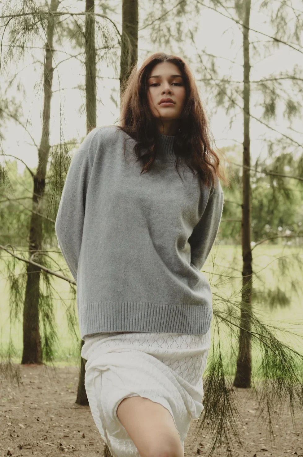 A young woman with dark hair stands outdoors in a forest, wearing a gray sweater and white shorts, looking at the camera with a neutral expression.