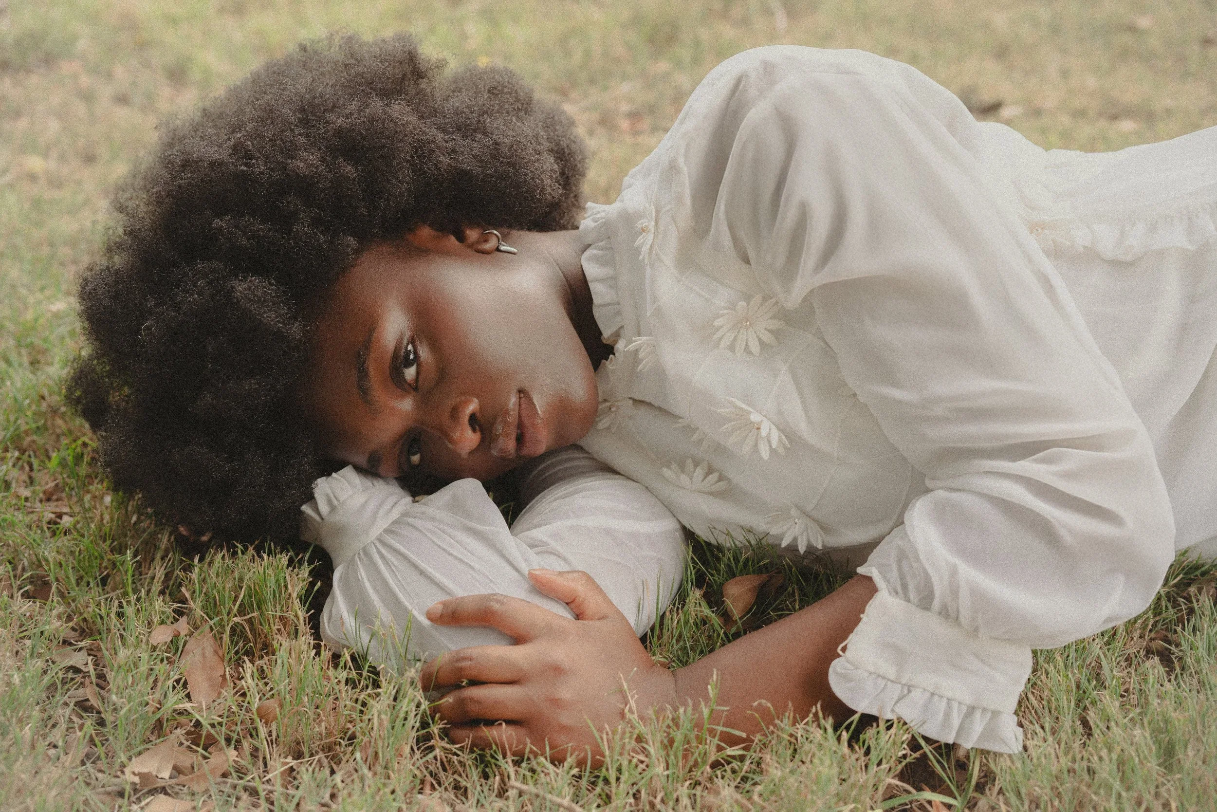 Young woman lying on grass, wearing a white blouse with flower embroidery, with dark curly hair, looking at the camera.