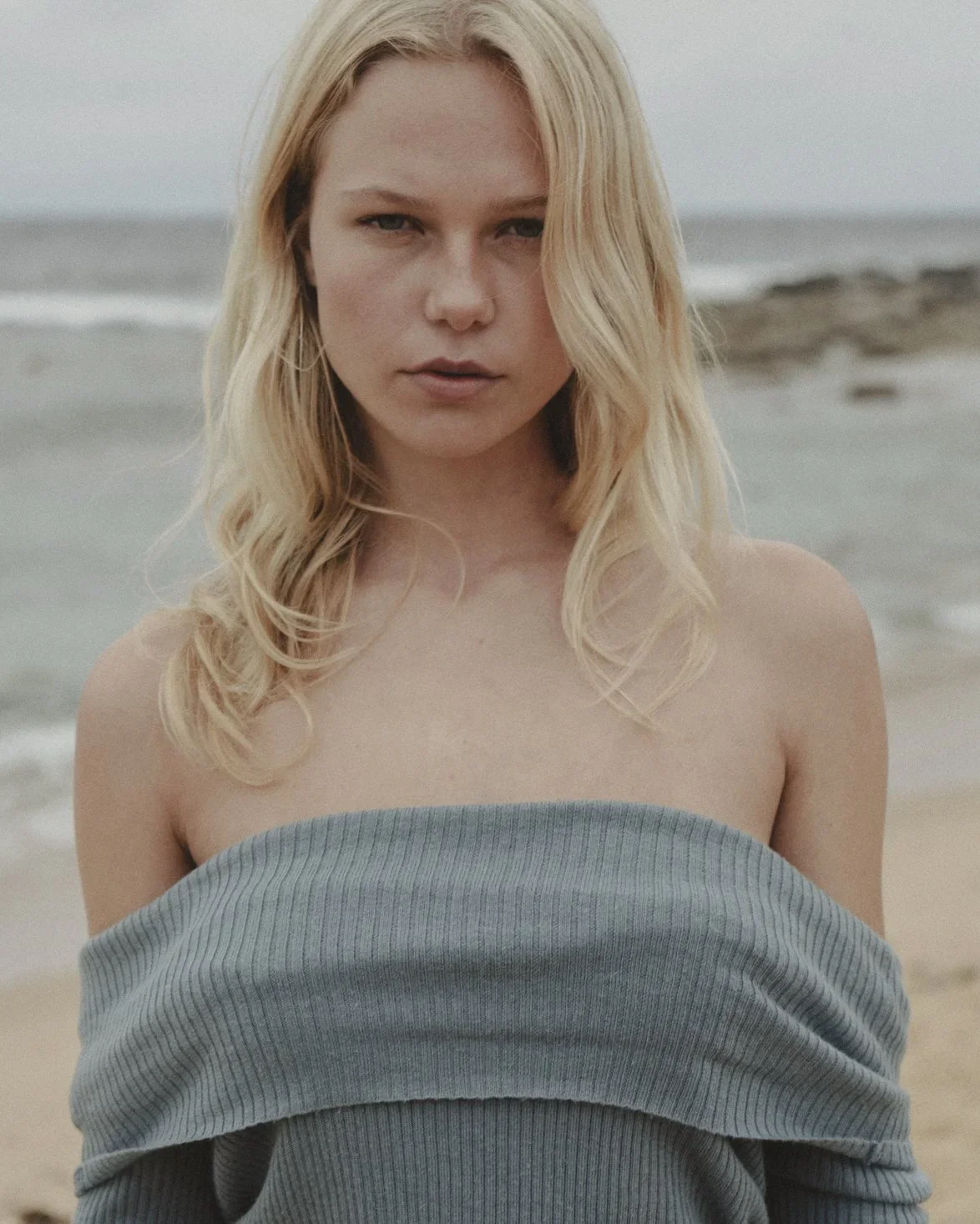 A woman with blonde hair standing on a beach, wearing a gray off-the-shoulder sweater, with the ocean and rocks in the background.