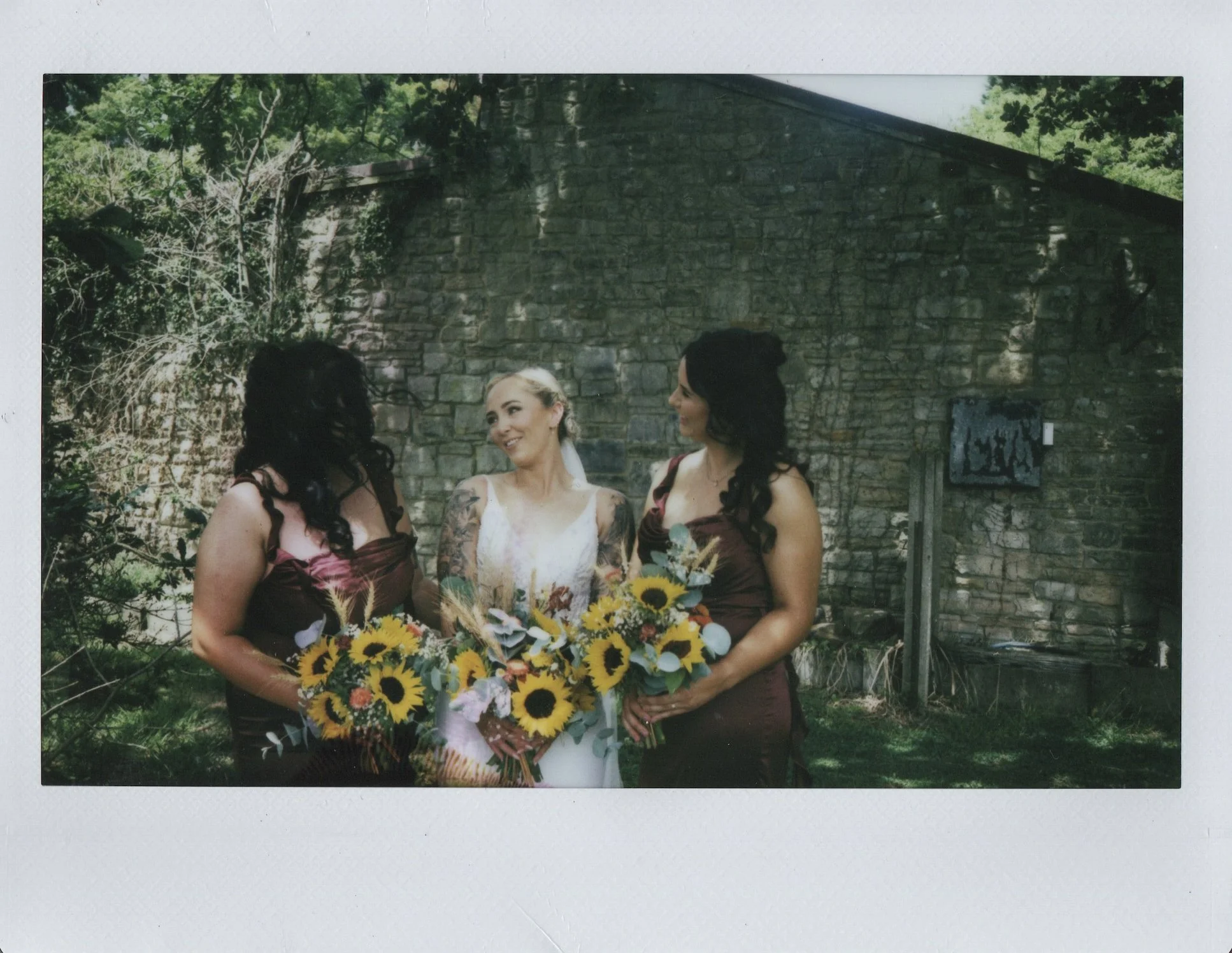 Three women, one in a white wedding dress and two in matching burgundy bridesmaid dresses, standing outdoors in front of a brick building. They are holding bouquets of sunflowers and other flowers, and are smiling at each other.