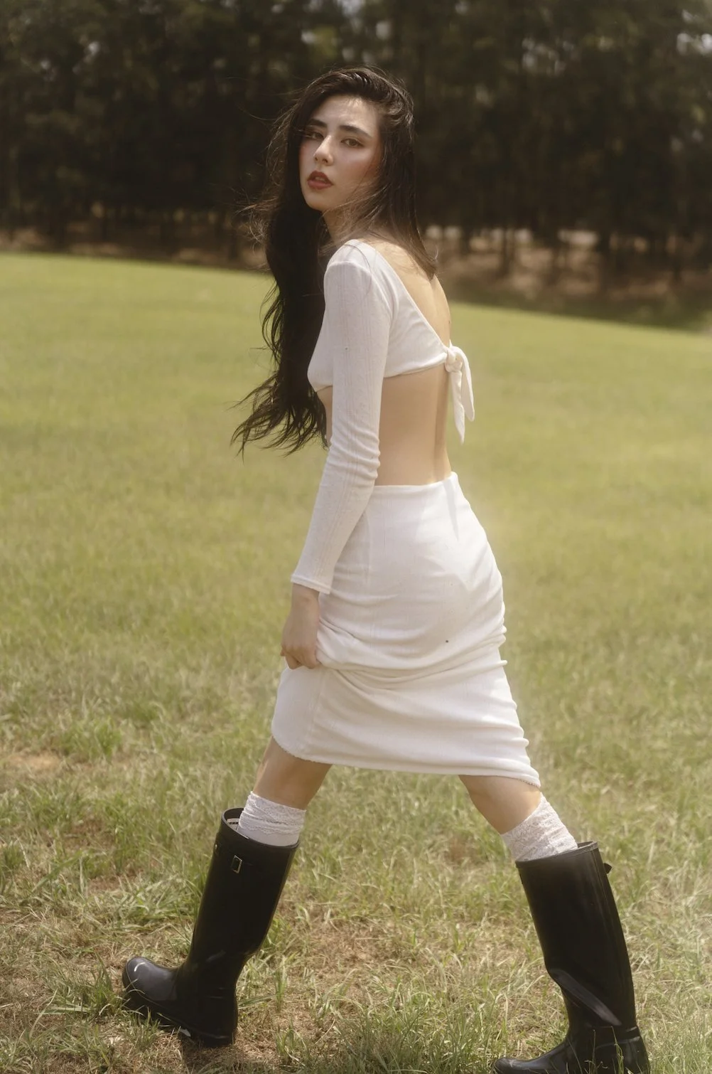 A young woman with long dark hair standing in a grassy field, wearing a white dress with a back bow, black rain boots, and white lace socks, looking at the camera with a serious expression.