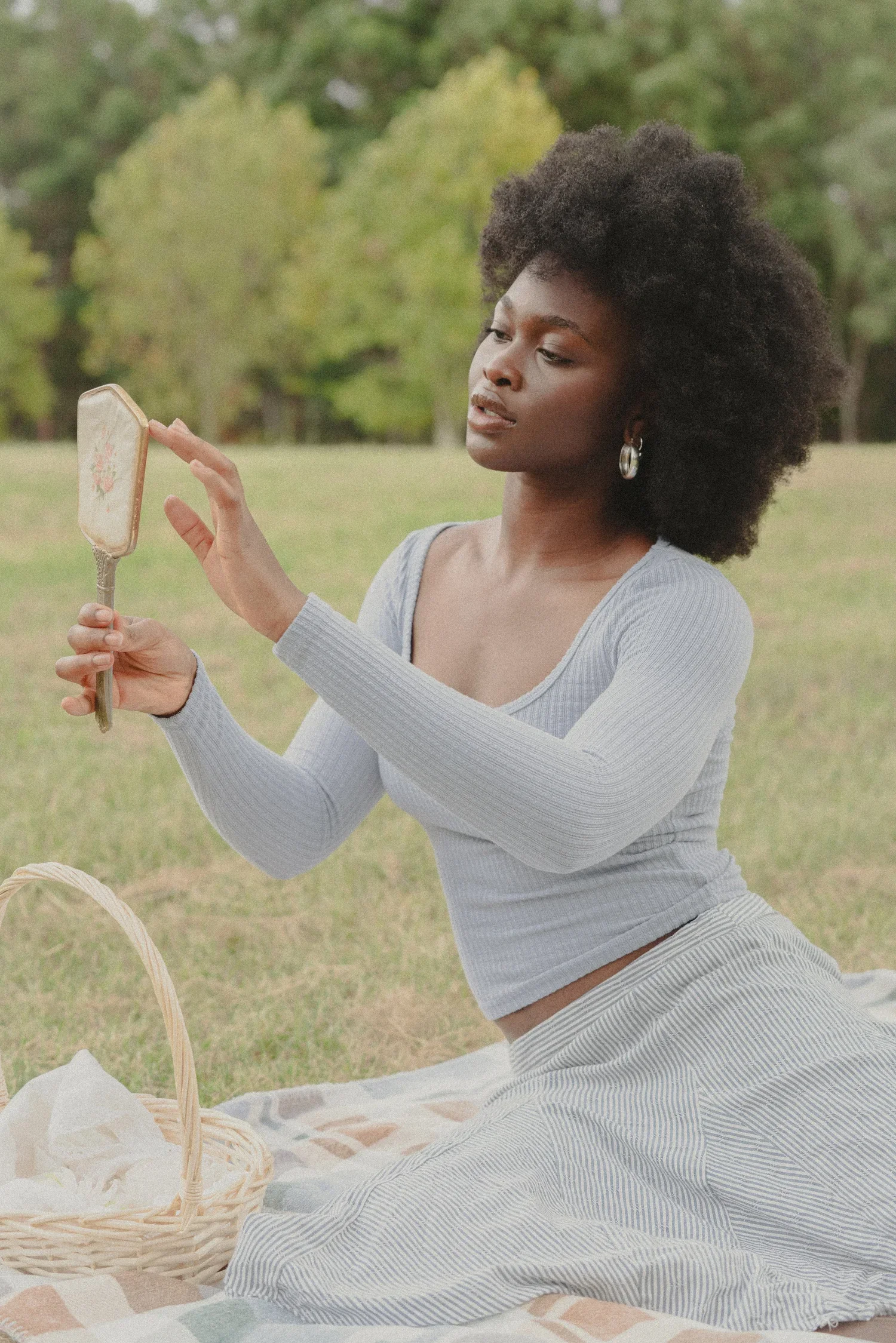 A woman sitting on a picnic blanket outdoors, holding a hand mirror, with a basket nearby, surrounded by trees.