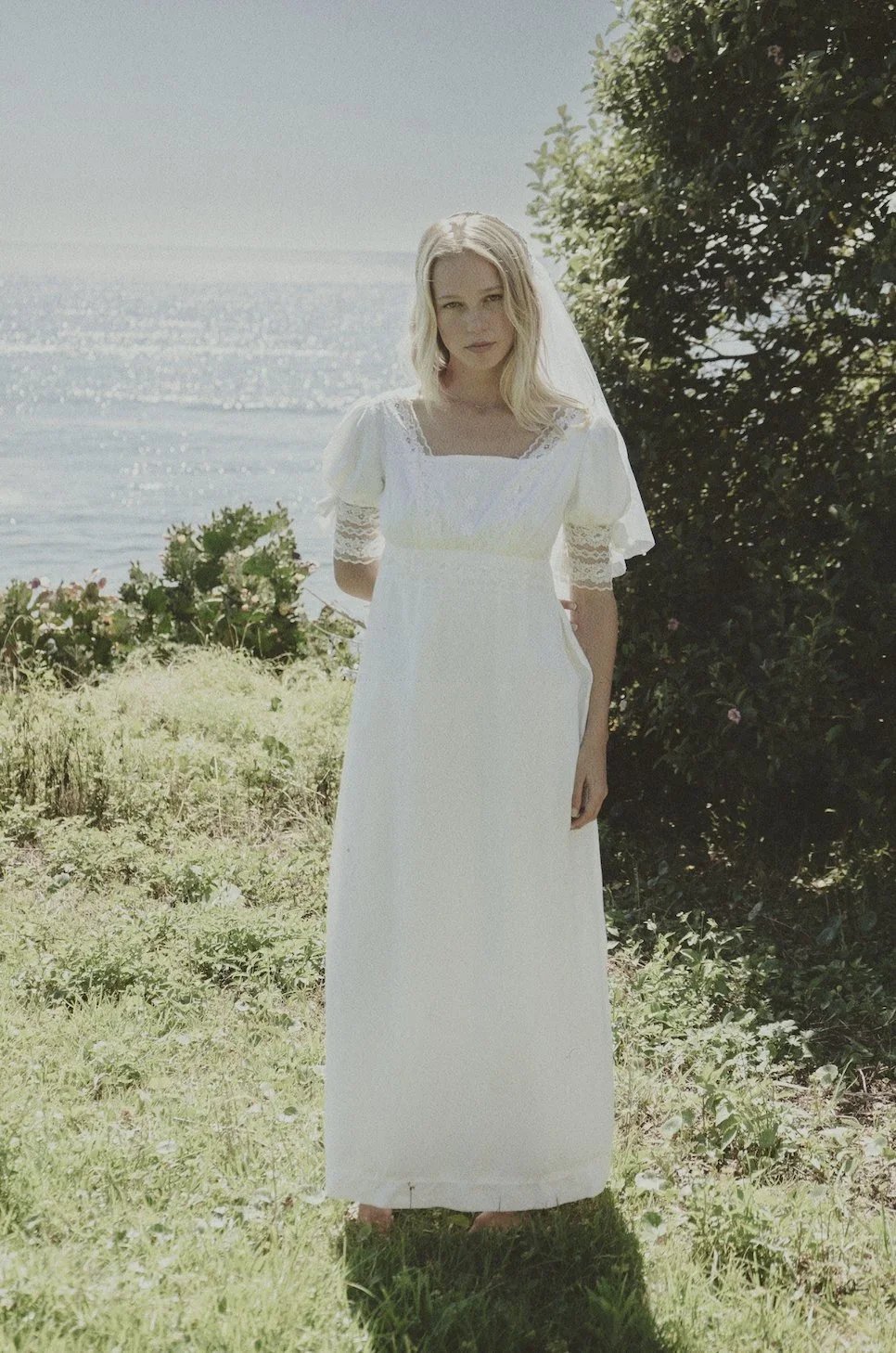 Woman in a white lace wedding dress standing outdoors on grass with a body of water and greenery in the background.