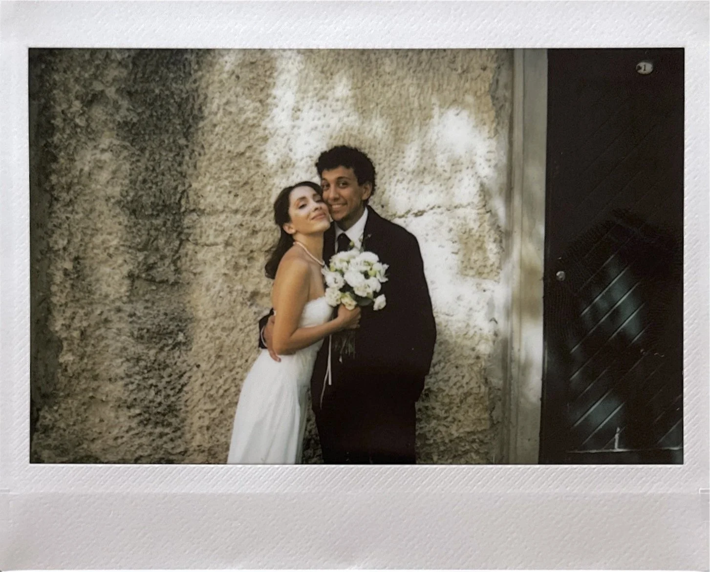 A newlywed couple standing close together, the woman in a white wedding dress holding a bouquet, and the man in a black suit, smiling, against a textured stone wall background.