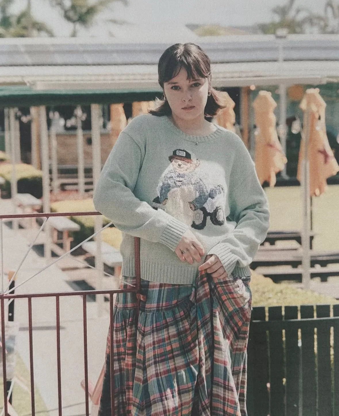 A young woman with short dark hair wearing a light gray sweater with a graphic of a bear dressed as a police officer, and baggy plaid pants, standing outdoors at a park or amusement area.