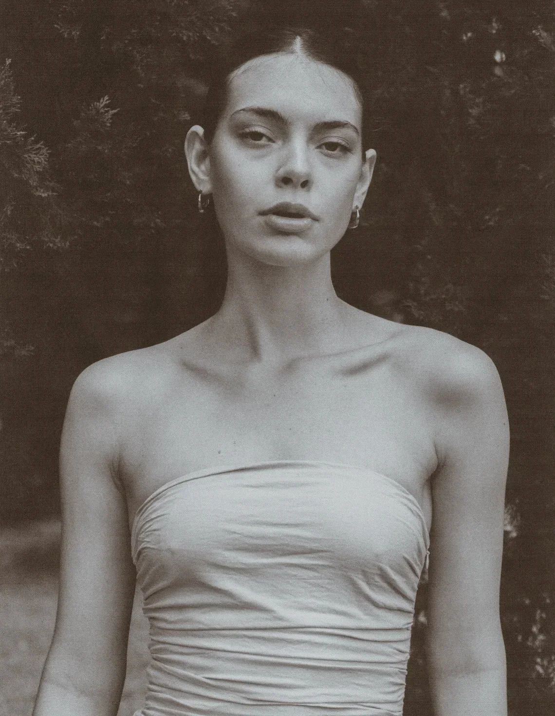 Black and white portrait of a woman with dark hair, wearing hoop earrings and a strapless top, standing outdoors with trees in the background.