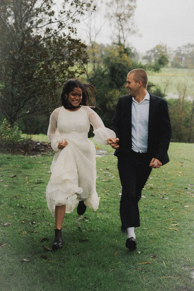 A happy couple running through a grassy park, woman in a white dress and man in a black suit, with trees and greenery in the background.