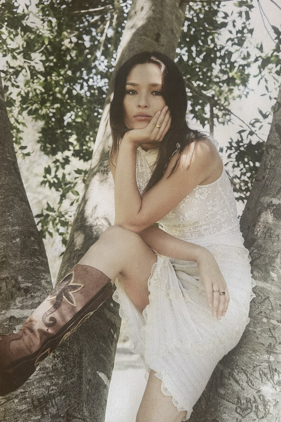 A woman with long dark hair sitting in a tree, resting her head on her hand, wearing a white lace dress and brown cowboy boots with floral embroidery.