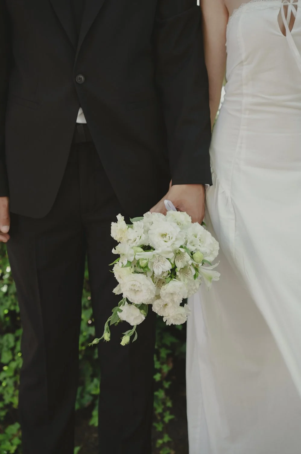A bride and groom holding hands, with the groom in a black suit and the bride in a white wedding dress, holding a white floral bouquet.