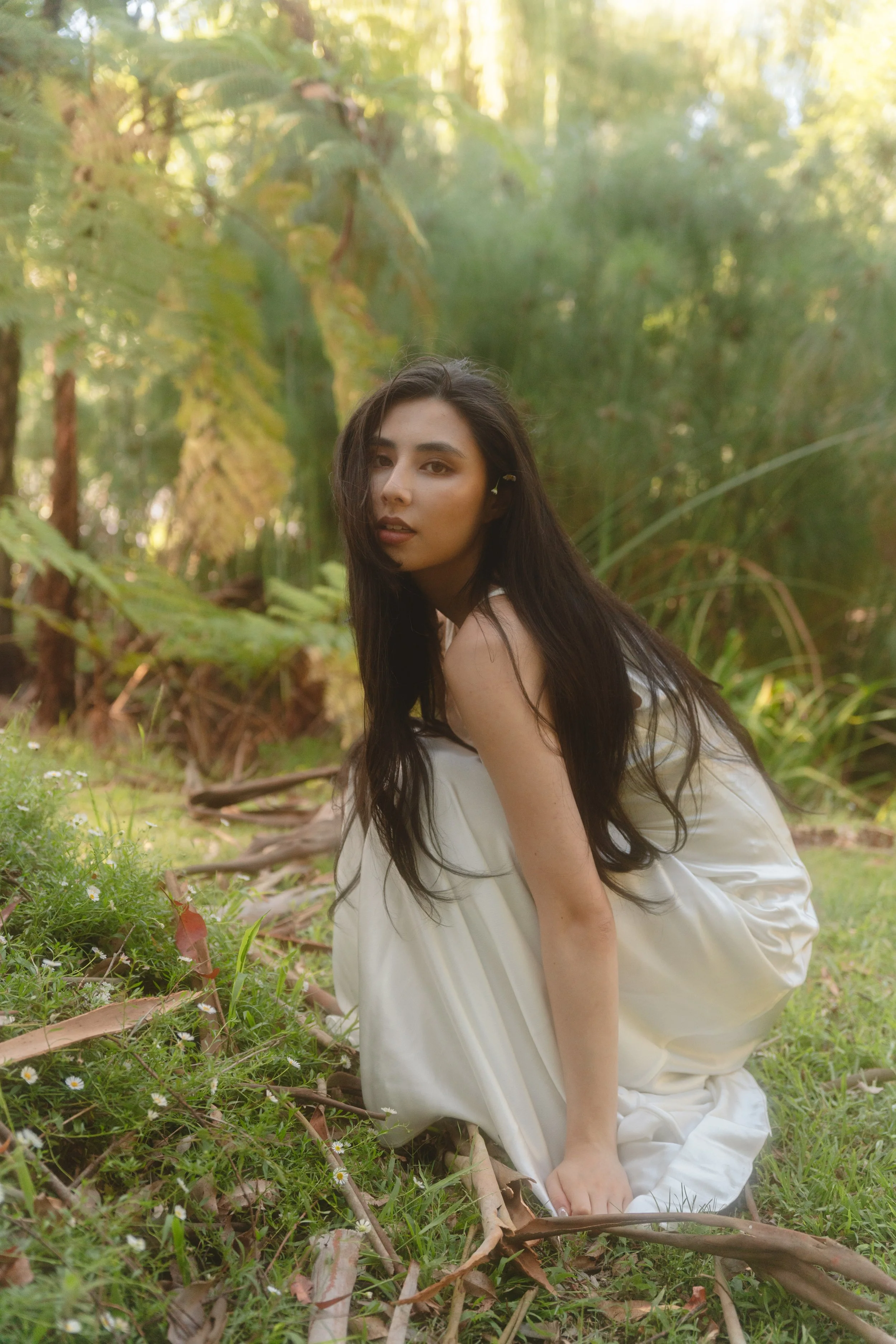 A woman crouching on the grass in a natural outdoor setting with green foliage and trees in the background.