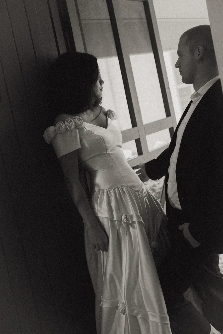 A black and white photo of a man and woman facing each other indoors. The woman is leaning against a wall, wearing a gown with decorative roses on the shoulder, and looking at the man. The man, dressed in a suit, is leaning against a window frame, holding the woman's hand, and looking at her.