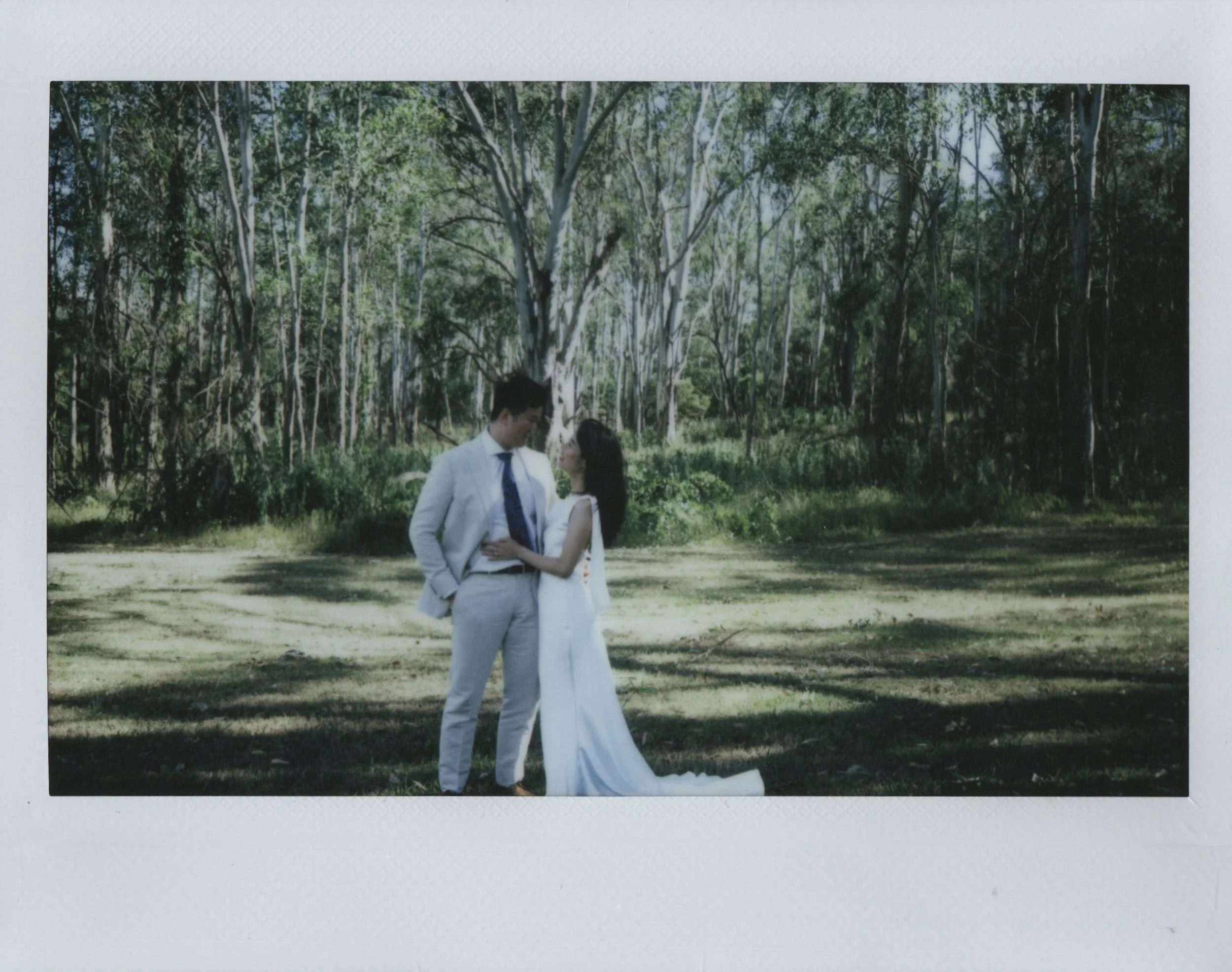 A bride and groom stand close together outdoors in a forest, gazing into each other's eyes. The groom is wearing a light-colored suit with a tie, and the bride is dressed in a white wedding gown. The background features tall trees and green foliage, with sunlight filtering through the leaves.