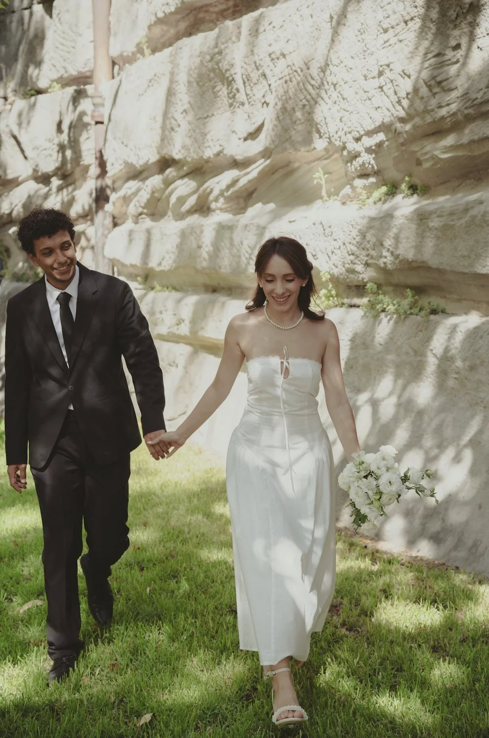 A bride and groom walking outdoors on grass, holding hands and smiling. The bride is wearing a white strapless wedding dress and holding a bouquet, while the groom is dressed in a black suit with a tie. They are walking near a large rock formation wi