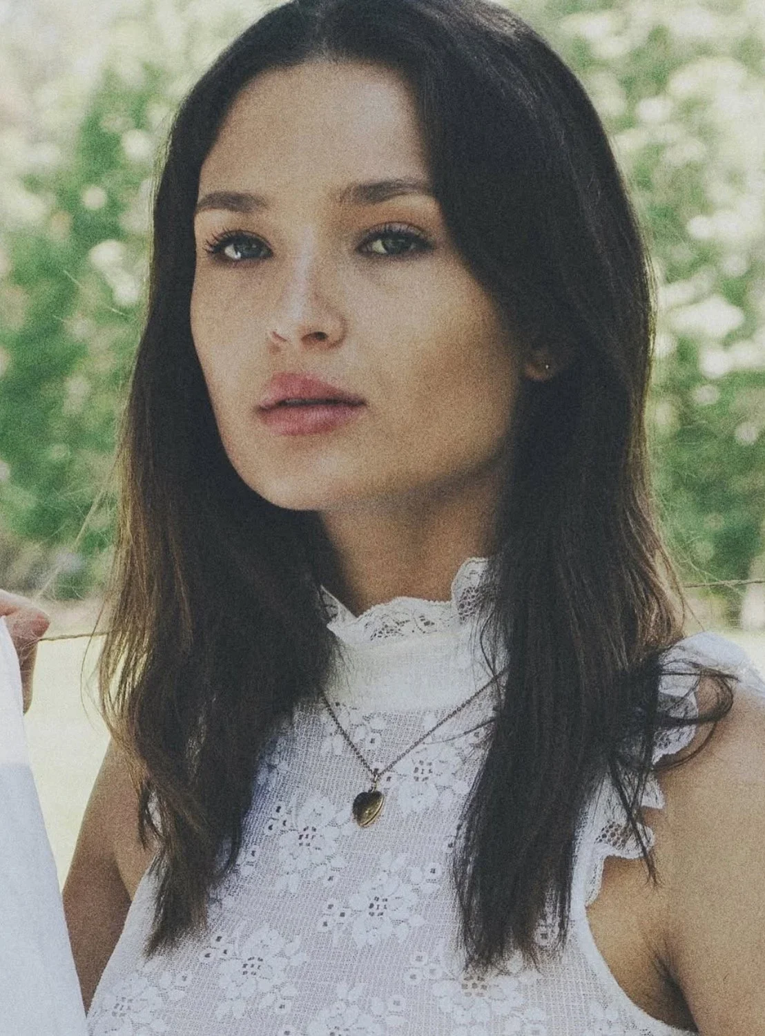 A woman with dark hair and light skin, wearing a white lace top with ruffled sleeves and a gold necklace in front of a blurred green outdoor background.