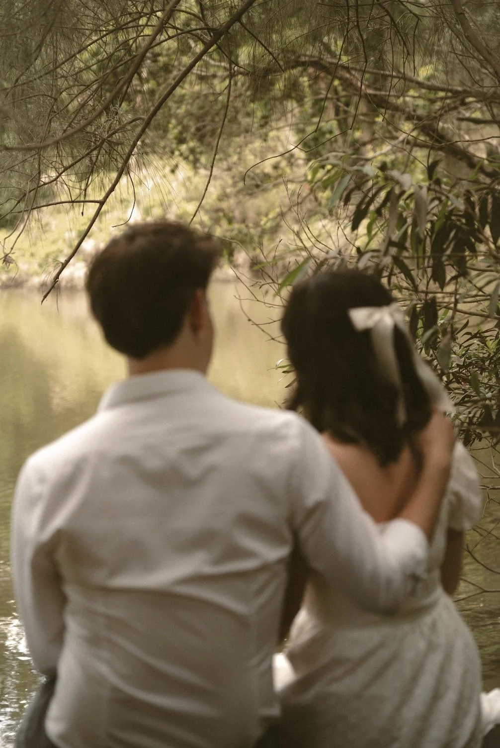 A man and woman sit together on a riverbank surrounded by trees, with the man gently holding the woman's head as they face away from the camera.