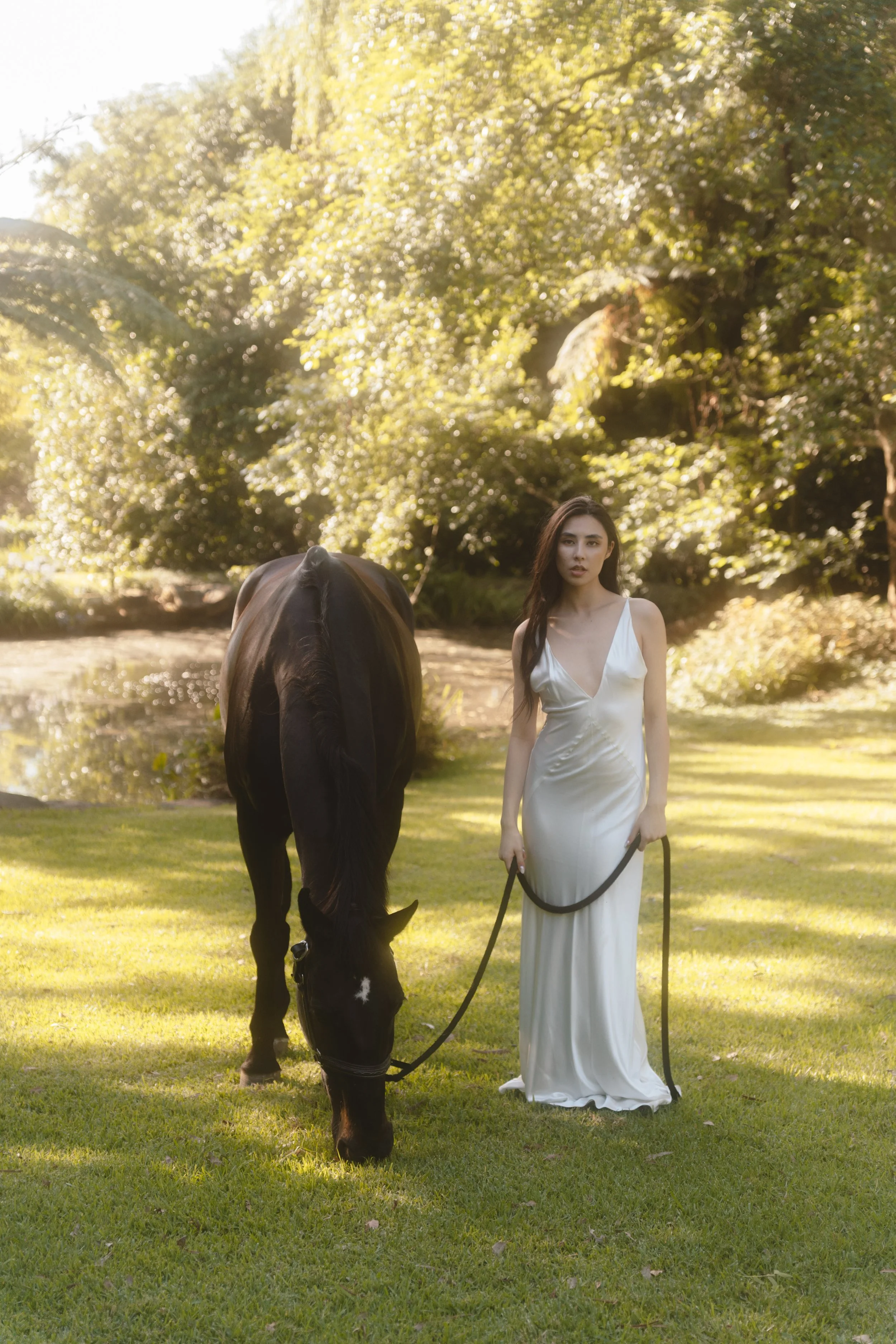 A woman in a white satin dress walking a black horse on a lush green lawn during daytime, with sunlight filtering through trees in the background.