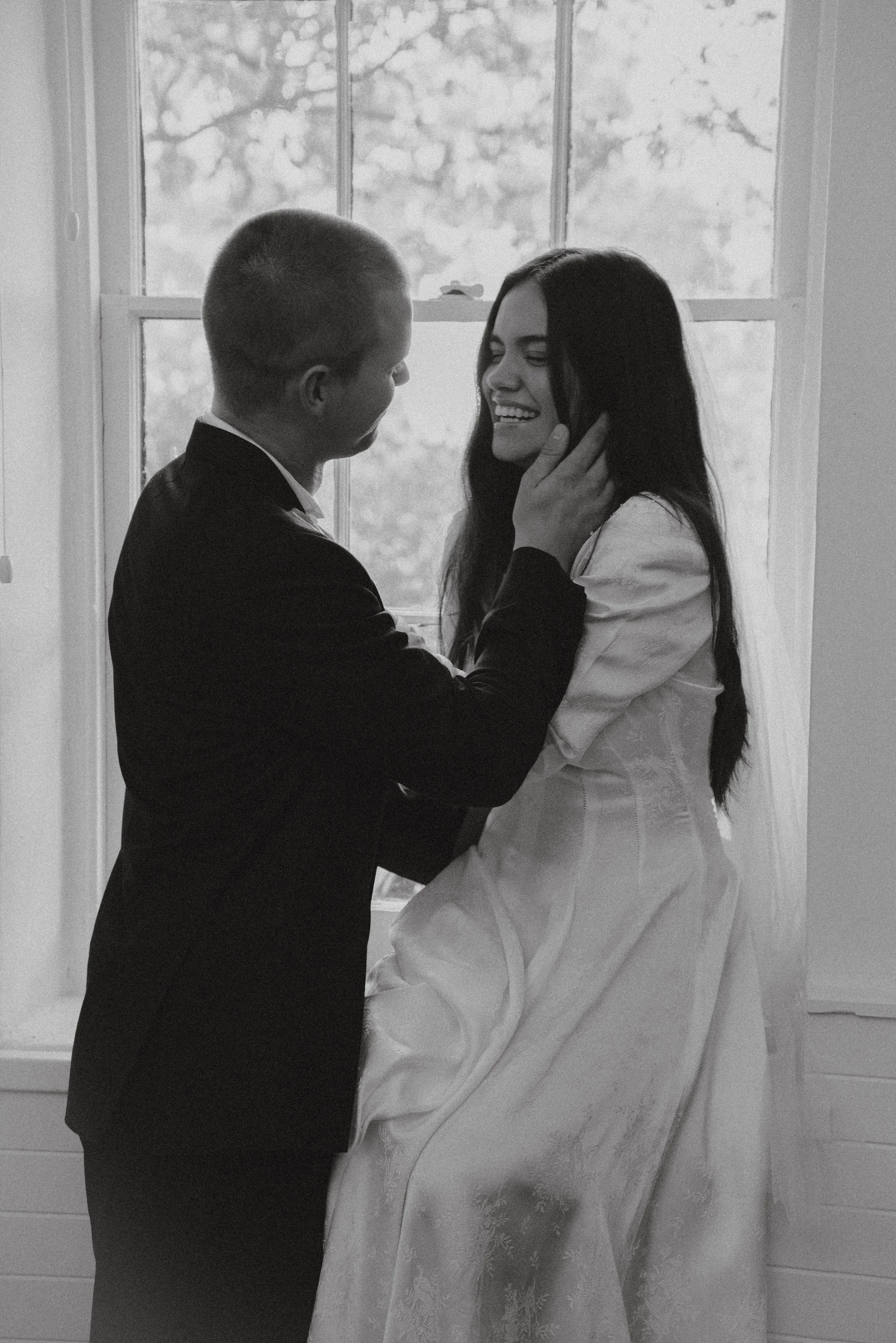 A black and white photo of a couple, with the man in a suit and the woman in a dress, holding each other's faces and smiling near a window.