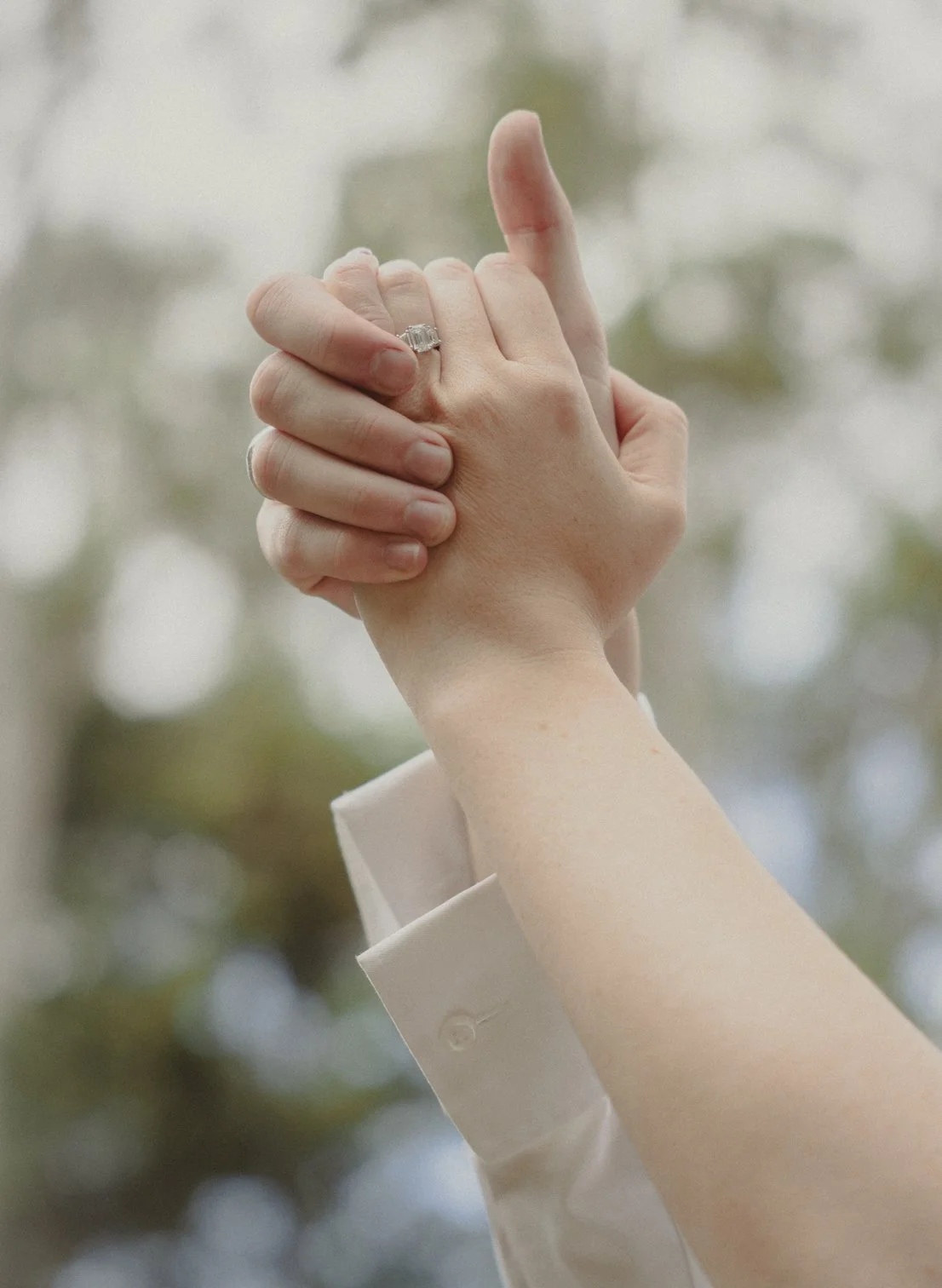 A close-up of two hands clasped together, with one person wearing a wedding ring, against a blurred background of trees and sky.