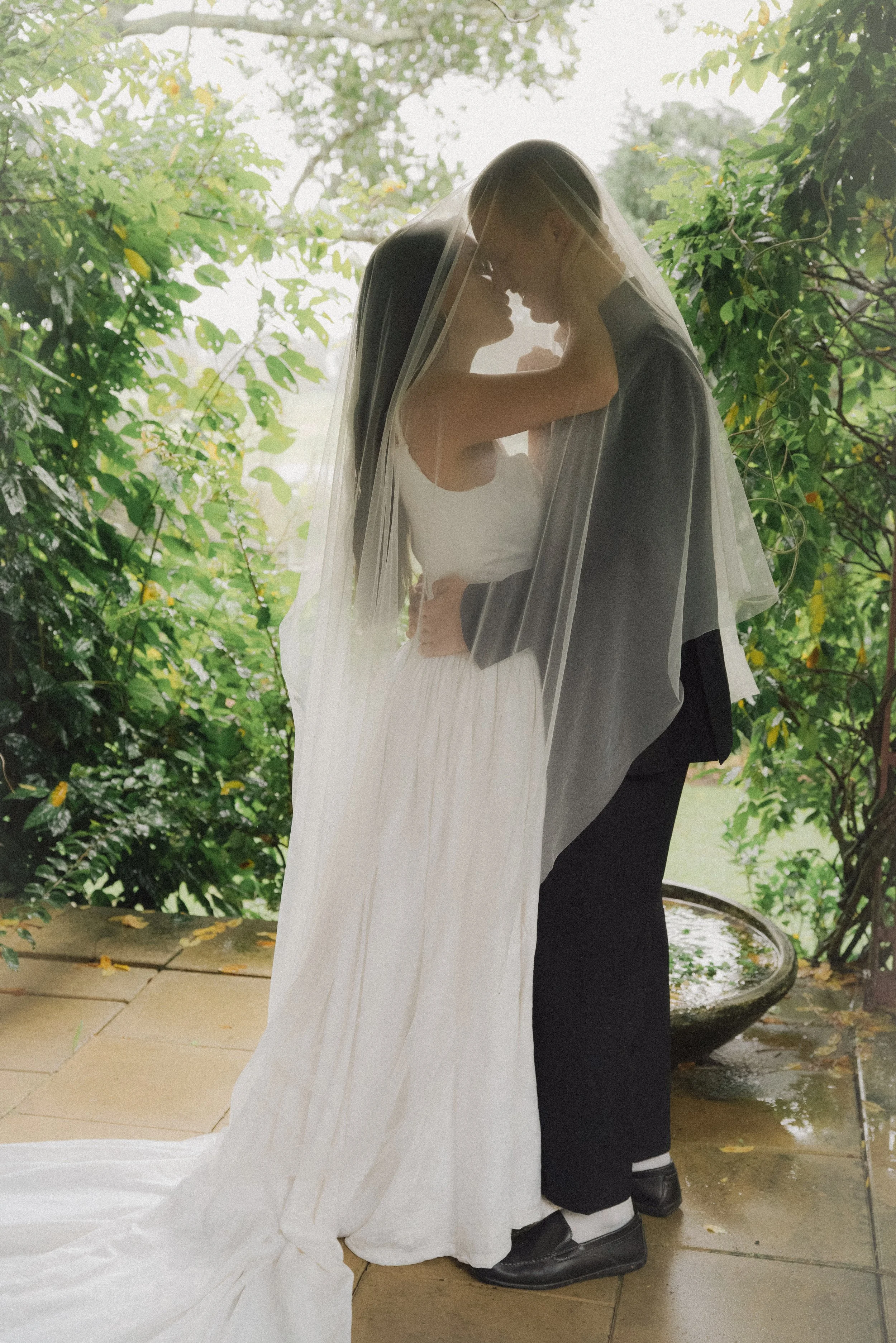 A bride and groom standing closely together under a sheer veil outdoors, surrounded by green foliage.