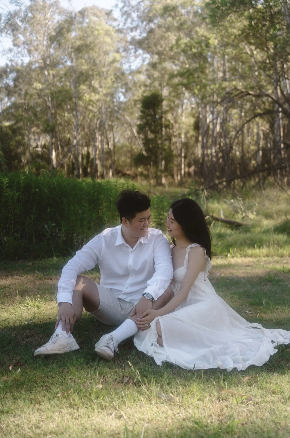 A couple sitting on the grass in a park, smiling and looking at each other, surrounded by trees with sunlight filtering through.