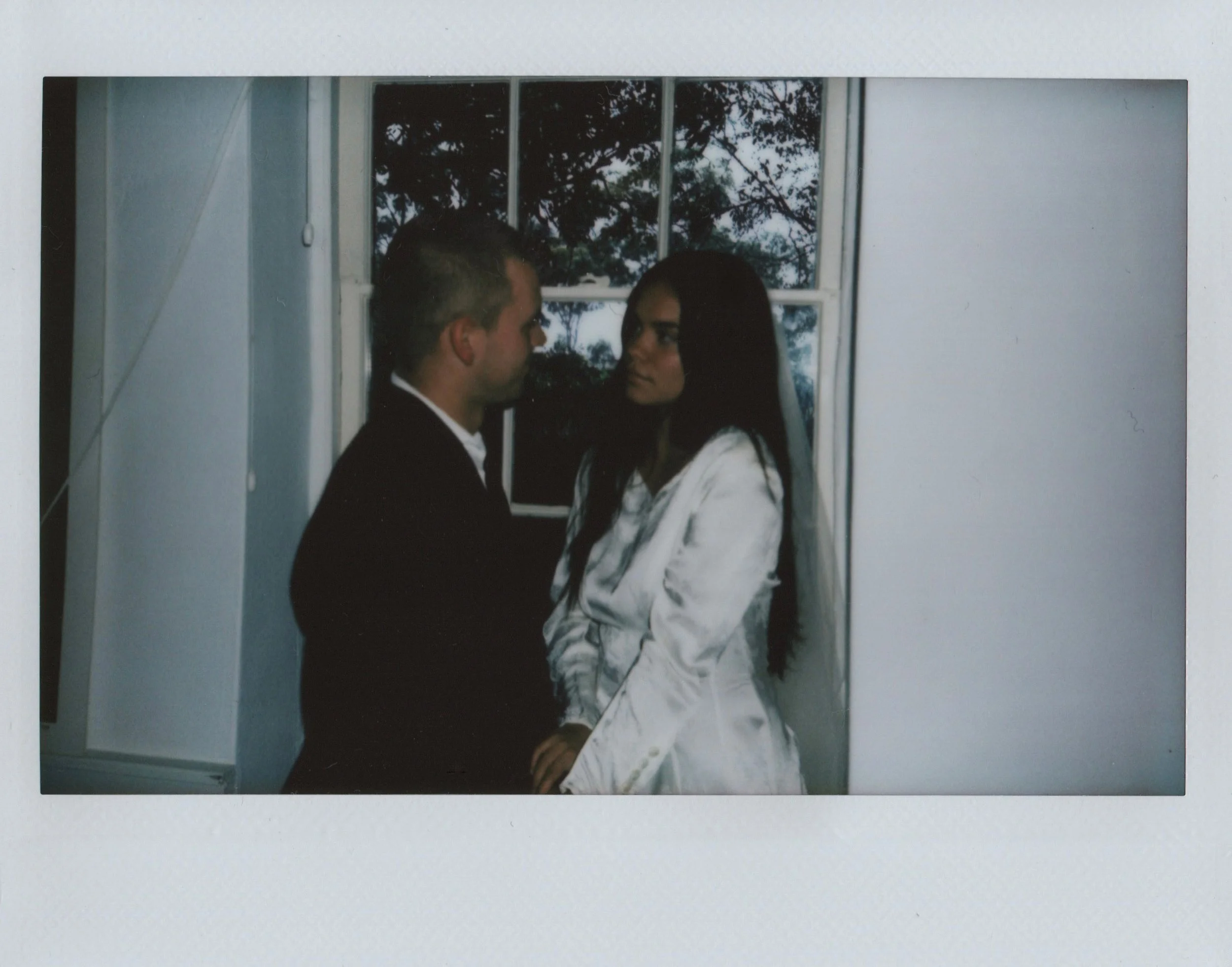 A man and woman face each other closely indoors, with a window showing trees outside behind them.