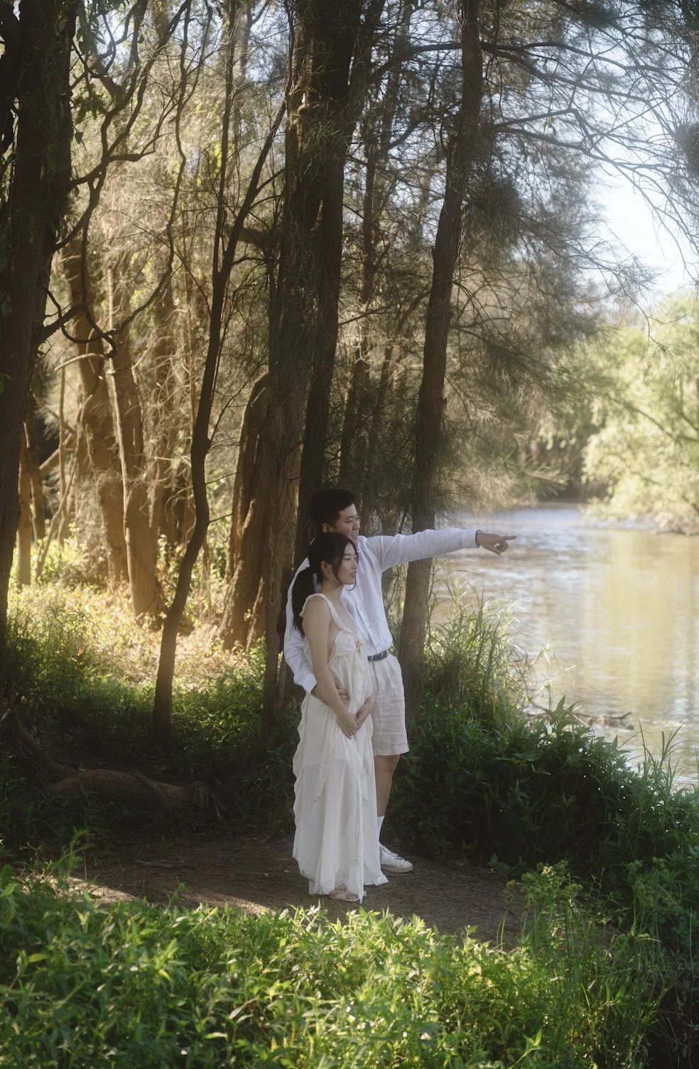 A couple stands by a river in a lush forest, with the man pointing towards something in the distance while the woman looks on.