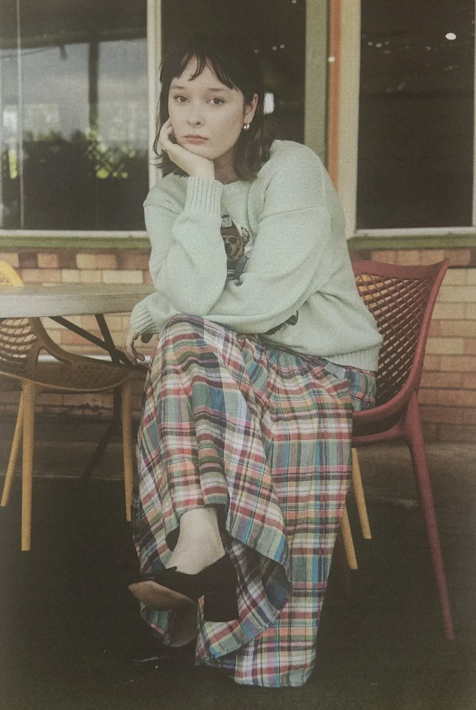 A young woman sits indoors at a table, resting her chin on her hand, wearing a light-colored sweater and a long, colorful plaid skirt. She has dark hair and looks directly at the camera. In the background, there are large windows, brick walls, and ch