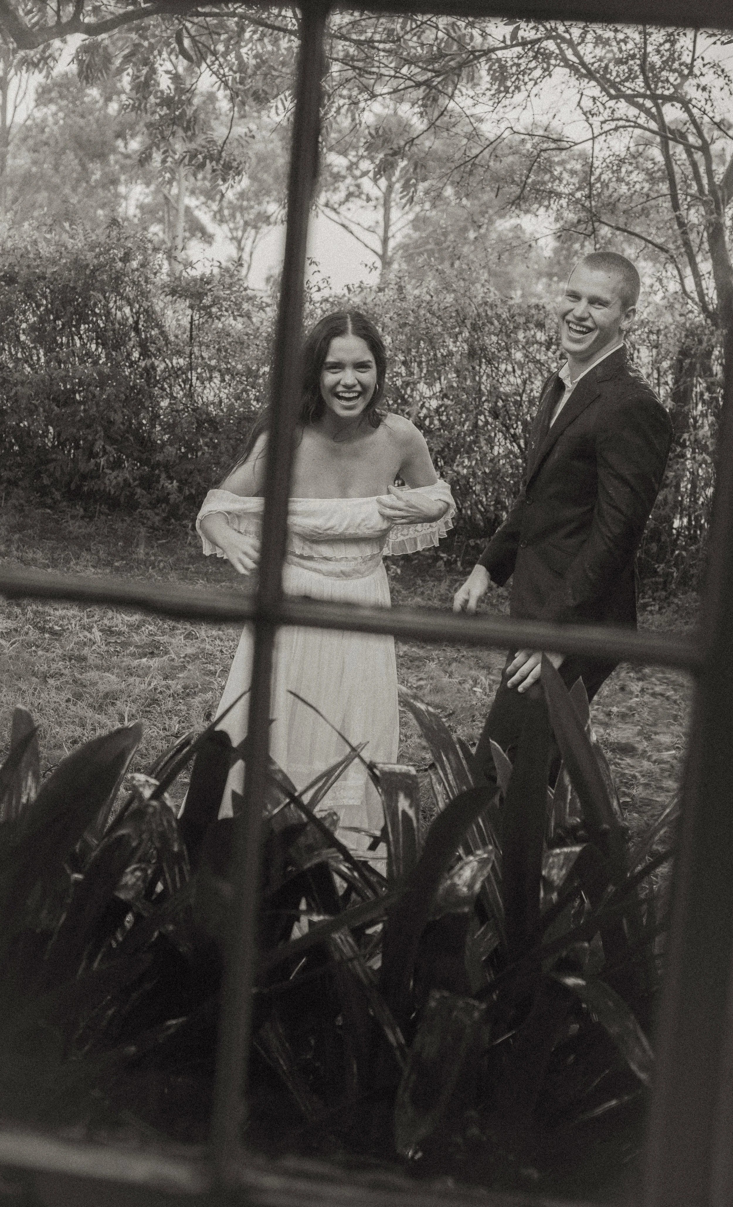 Black and white photograph of a smiling young woman in an off-the-shoulder dress and a young man in a suit standing outdoors near bushes and trees, seen through a window with grill bars.