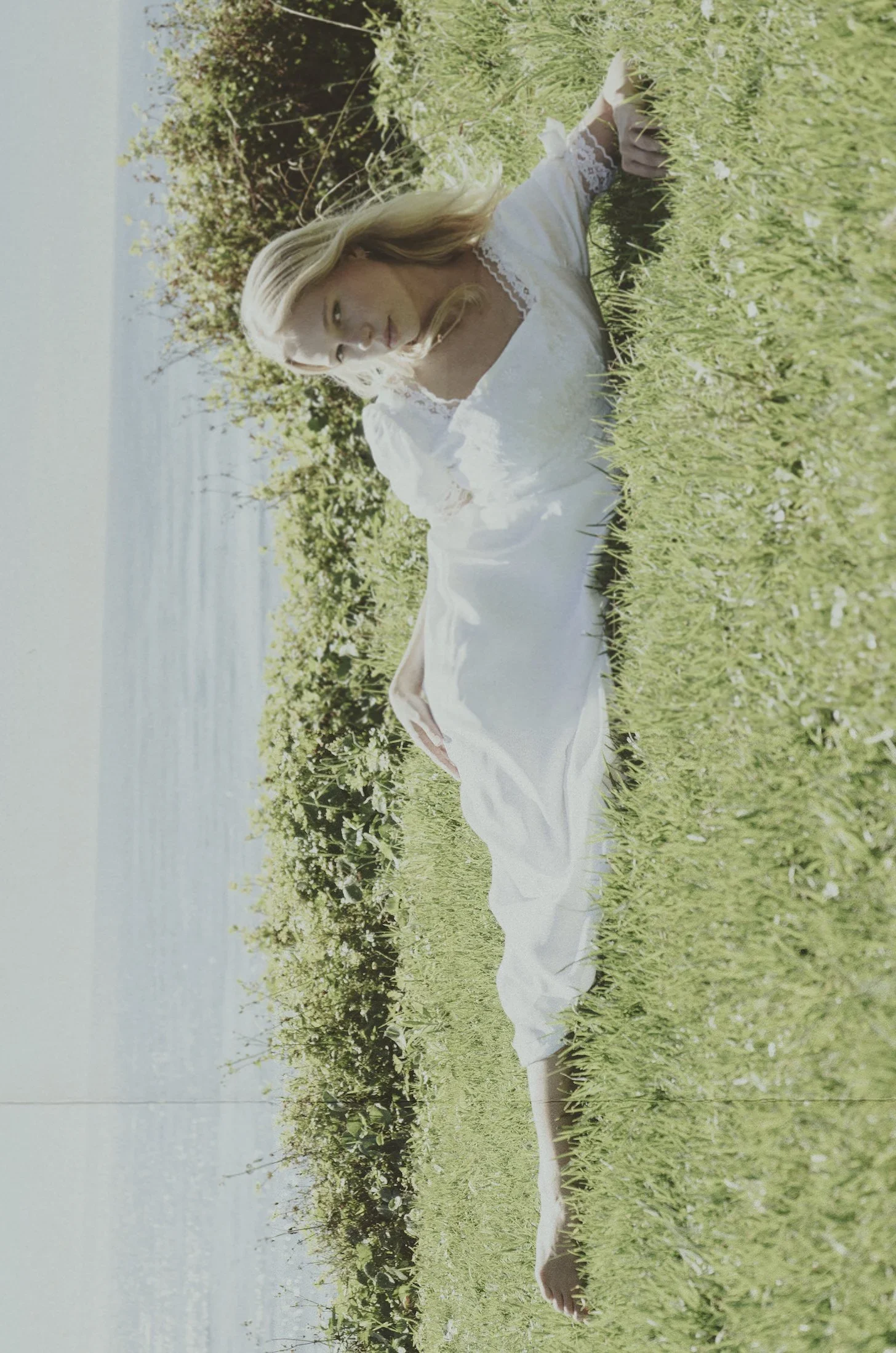 A woman in a white dress is lying on green grass near a body of water, with greenery and trees in the background. She is looking to her side, with her head resting on her arm.