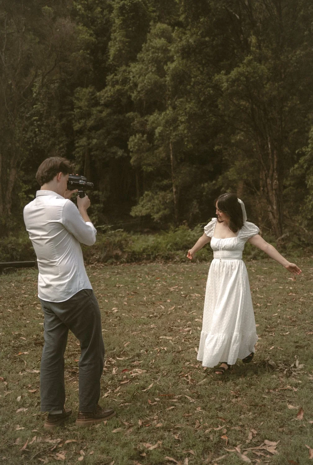 A woman in a white dress posing in a wooded area while a man in a white shirt takes her photo with a camera.