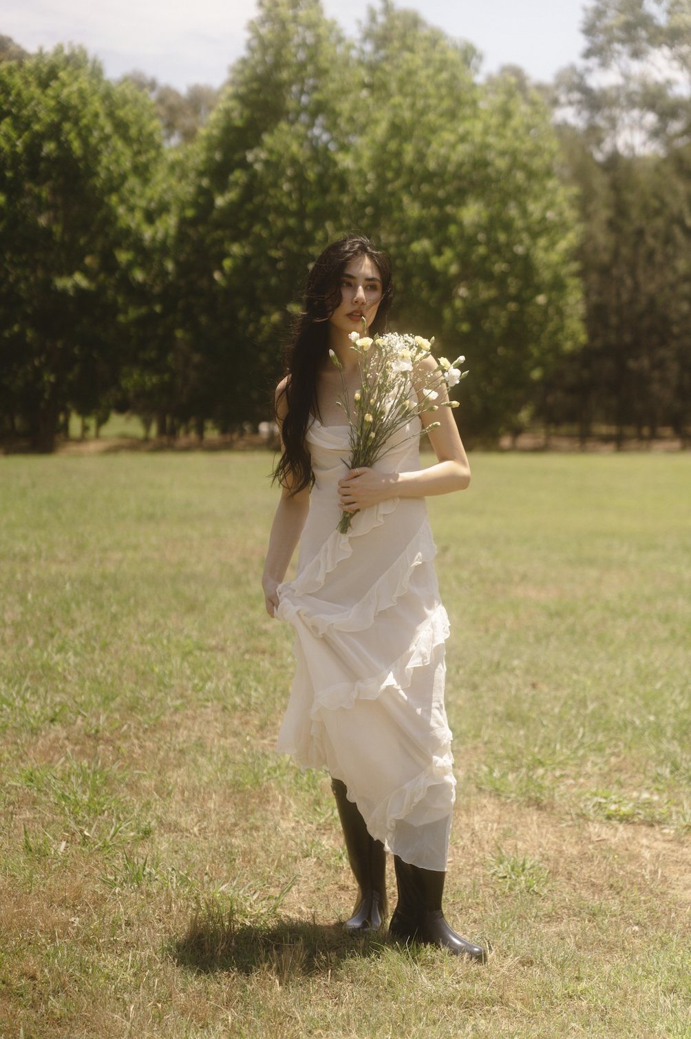 A woman with long dark hair wearing a white dress and black rain boots standing outdoors on a grassy field holding a bouquet of white flowers, with trees in the background.
