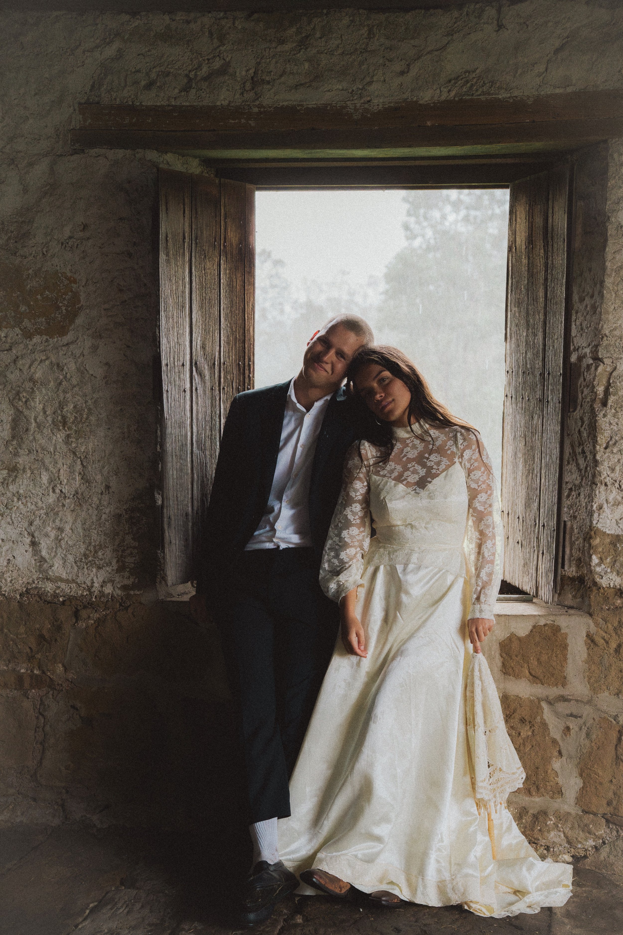 A couple on their wedding day sitting next to an open window with wooden shutters, stone wall background, rain outside, groom in black suit and white shirt, bride in cream gown with lace sleeves.