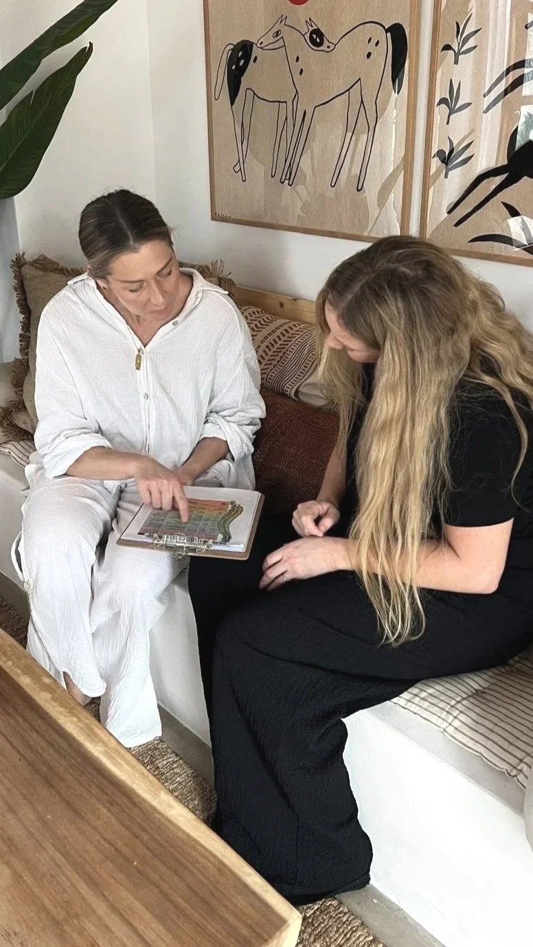 Two women sitting on a bed, examining a tablet together, with a wall art of two abstract horses and plant leaves in the background.