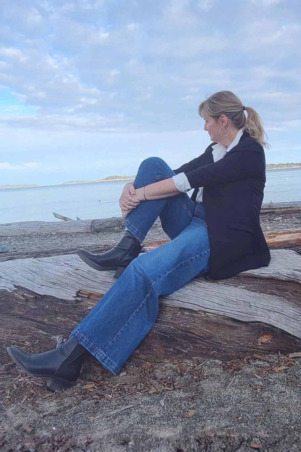 Sherri Kozubal, certified medical intuitive and hypnotherapist sitting on a piece of driftwood by the ocean