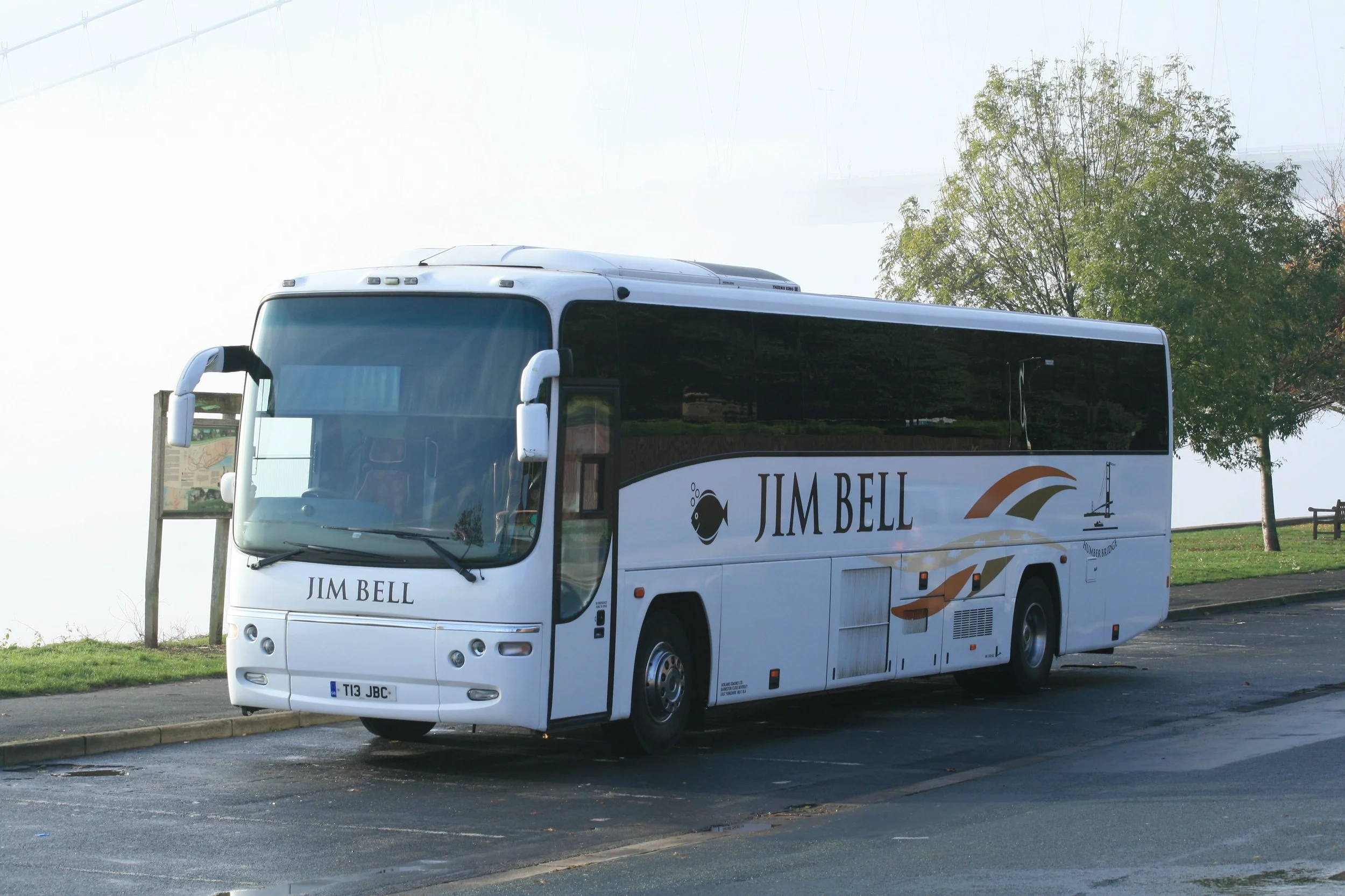 White tour bus with Jim Bell logo parked on street near park with grass, trees, and bench