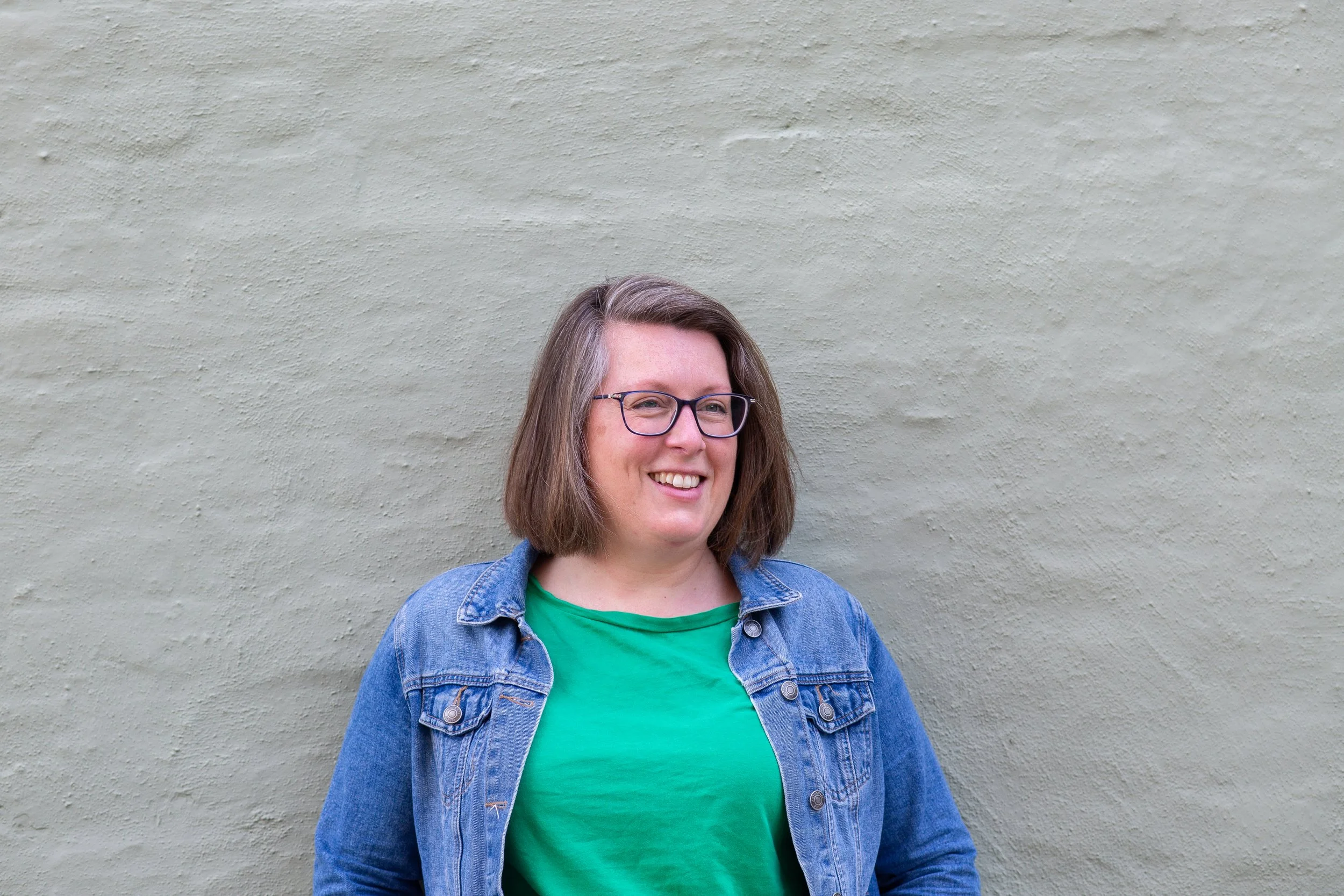 A woman with shoulder-length brown hair, glasses, wearing a green shirt and denim jacket, smiling while standing against a light grey textured wall.