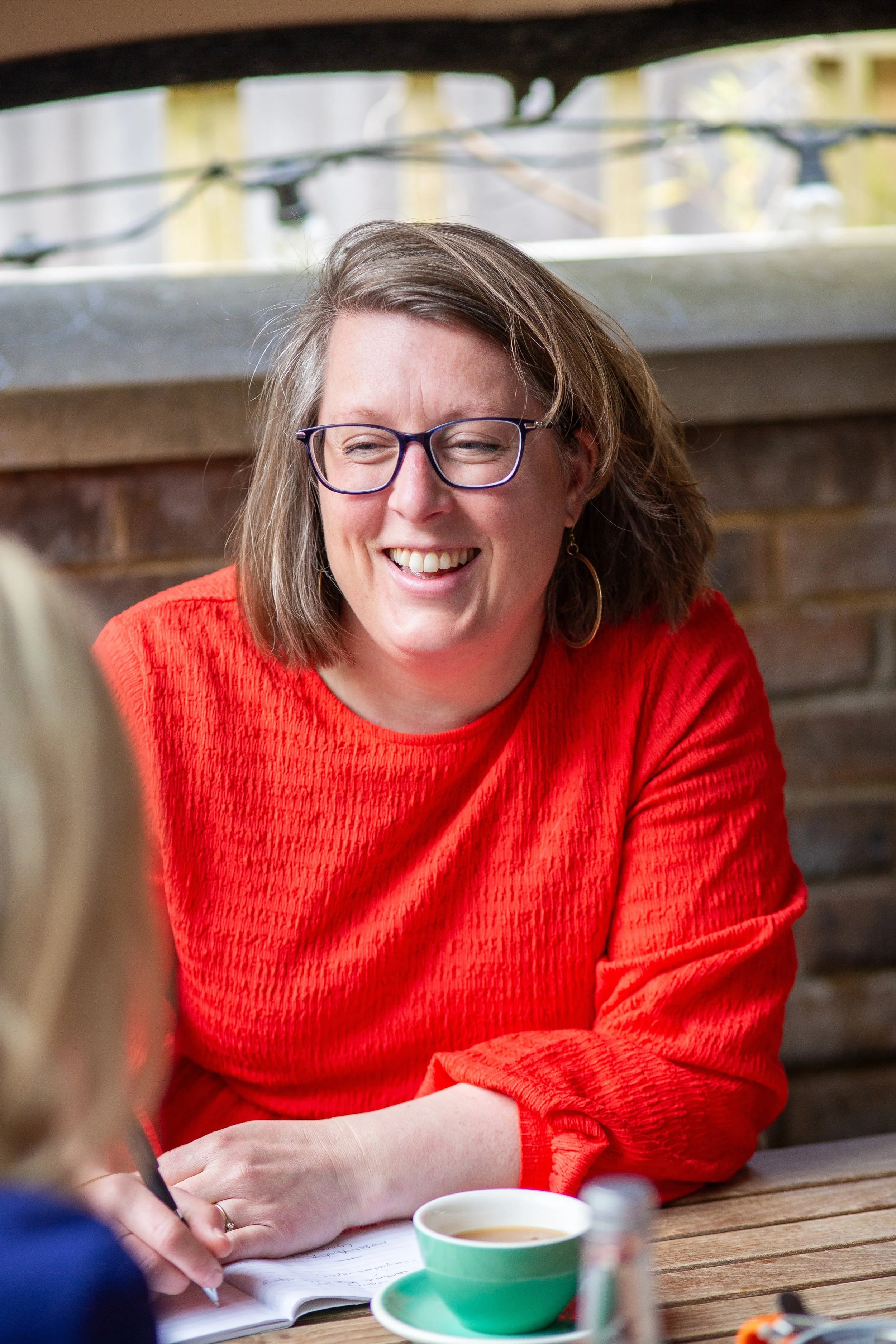 Picture of Anna Foster - with shoulder-length brown hair, glasses, and earrings, smiling while sitting at a wooden table outdoors, wearing a red sweater, with a cup of tea and a notebook in front of her, talking informally with a client