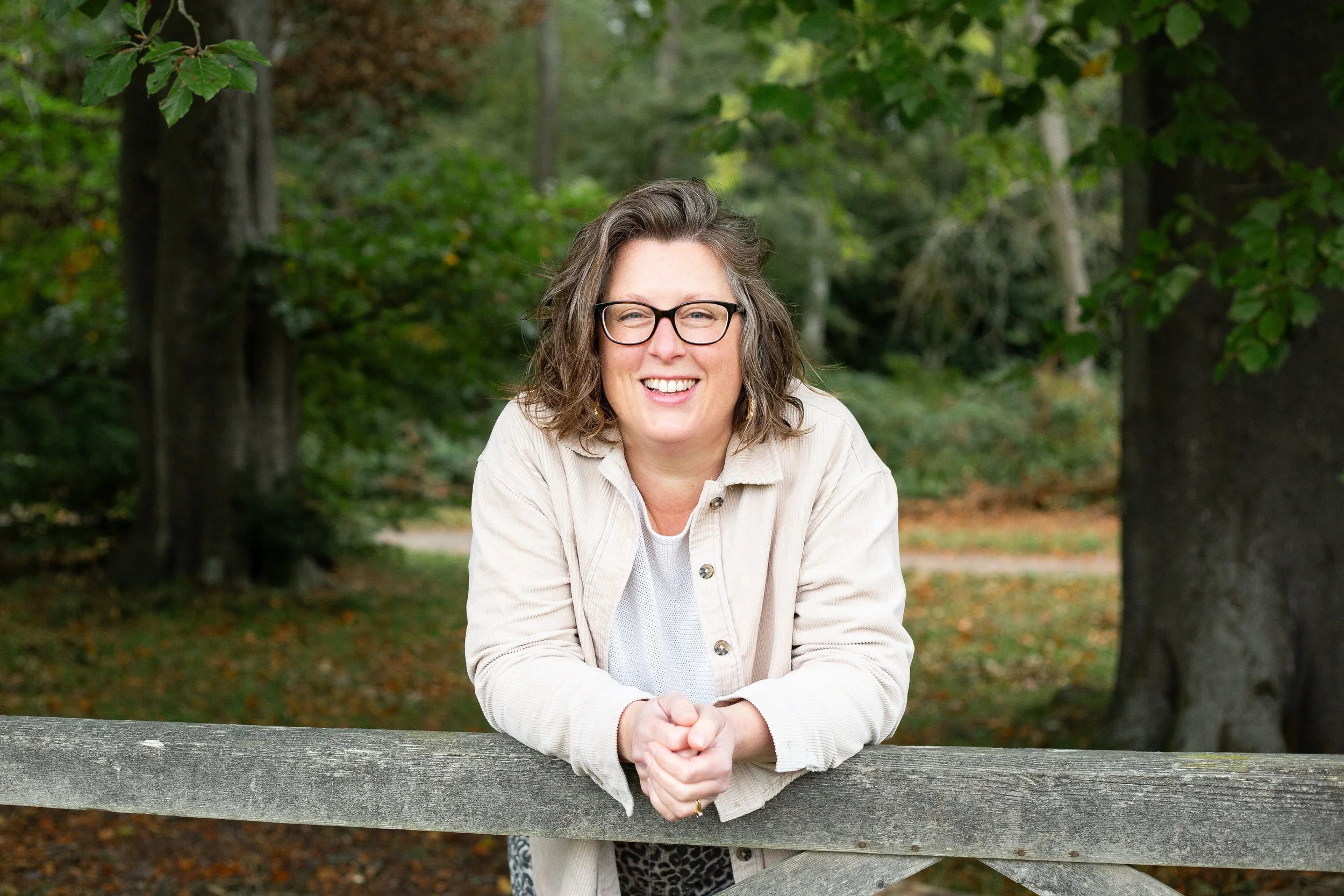 Picture of Anna Foster - with glasses and curly hair smiling outdoors, leaning on a wooden fence in Ashridge forest, surrounded by trees