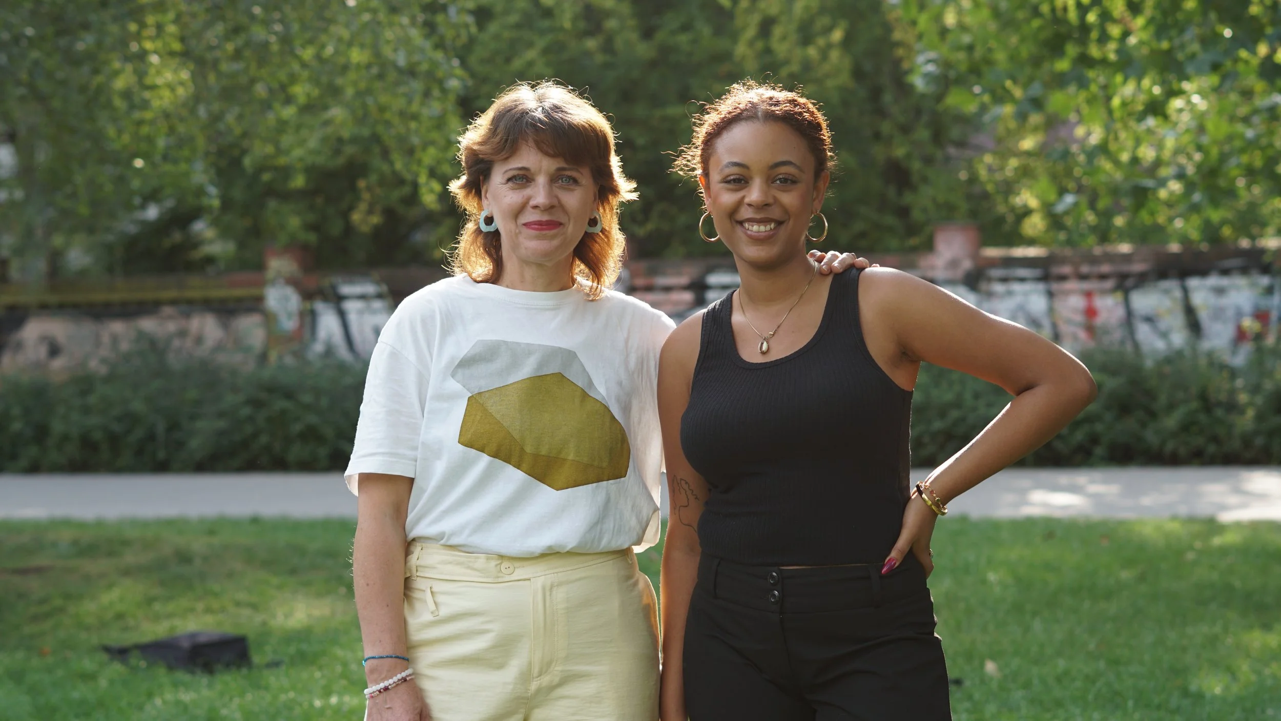 Zwei Frauen stehen nebeneinander in einem Park, lächelnd in die Kamera schauend, umgeben von grünen Bäumen und Wiesen, mit einer Brücke im Hintergrund.